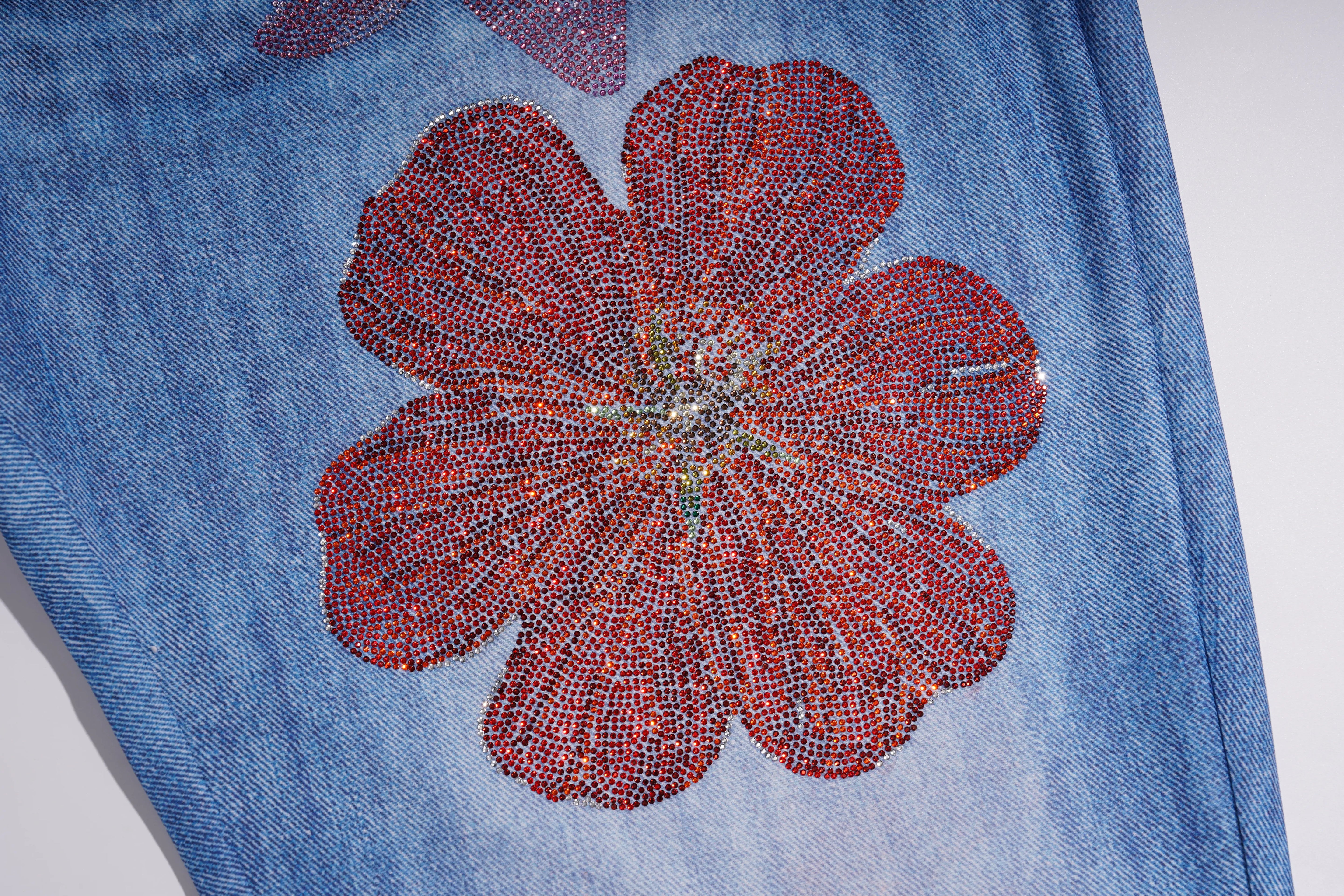 Close-up of a blue denim garment featuring a large red beaded flower applique on the front, with a small multicolor floral motif partially visible above it. TCH Floral Rhinestone Wide Leg Denim Jeans