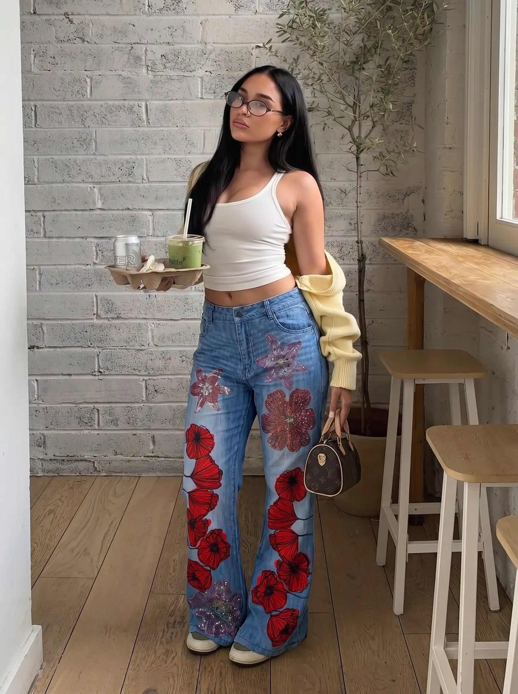 A woman stands in a cafe-like interior wearing blue wide-leg jeans with large red floral prints, a white ribbed tank top, and a pale yellow cardigan draped off her shoulder. She holds a tray with drinks and carries a small dark handbag. TCH Floral Rhinestone Wide Leg Denim Jeans
