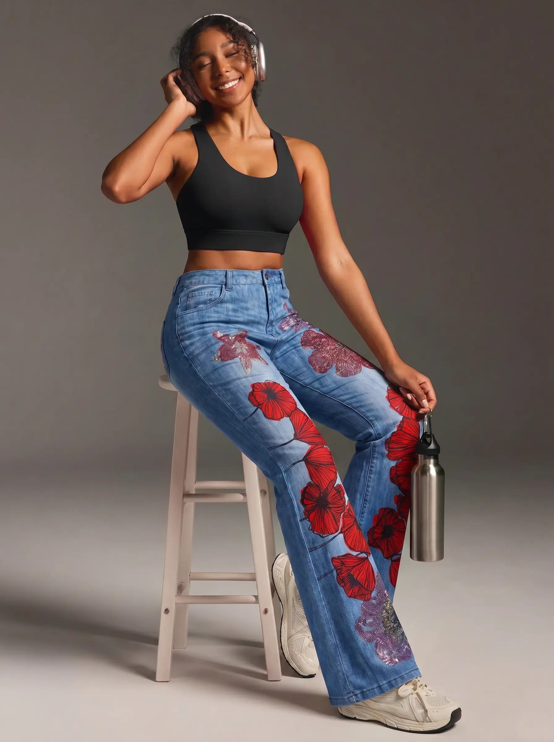 A woman sitting on a white stool wearing blue jeans with a red poppy floral print, a black sports bra, and white sneakers, holding a silver water bottle against a gray studio background. TCH Floral Rhinestone Wide Leg Denim Jeans