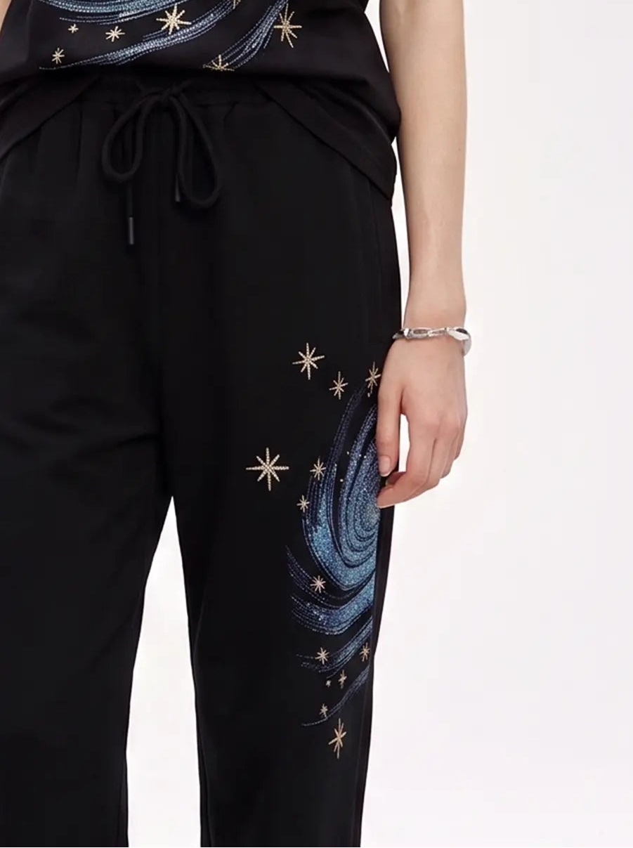 A woman modeling a black short-sleeve t-shirt and matching black drawstring pants set, both featuring a blue spiral galaxy print and scattered gold star details, against a plain light gray studio background. TCH Galaxy Rhinestone Wide Leg Sweatpants