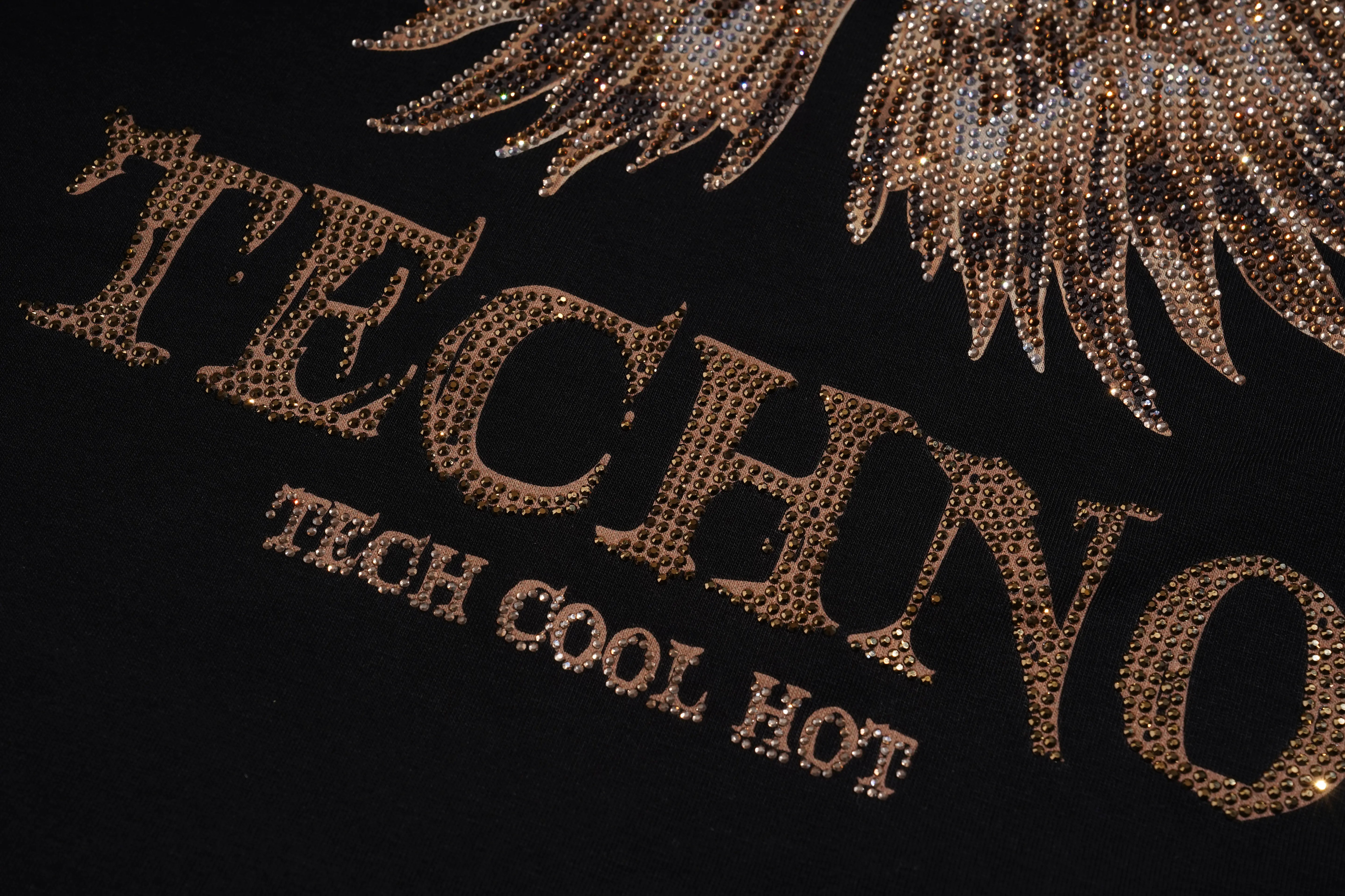 Close-up of a black garment featuring large metallic gold rhinestone-style lettering that reads 'TECHNO' with smaller text 'TECH COOL HOT' beneath it, plus a matching gold wing design at the top. TCH Golden Wing Techno Oversized Graphic T-Shirt