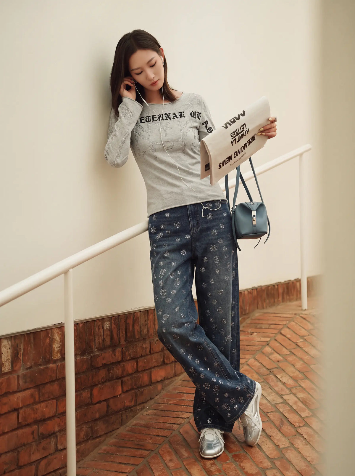 A woman stands on a brick walkway against a cream wall, wearing a light gray long-sleeve graphic sweatshirt, blue jeans with a small white monogram pattern, and white sneakers. She is holding a newspaper and carrying a small blue shoulder bag. TCH Gothic Rhinestone Lace Pattern Flared Sleeve Long Sleeve Top