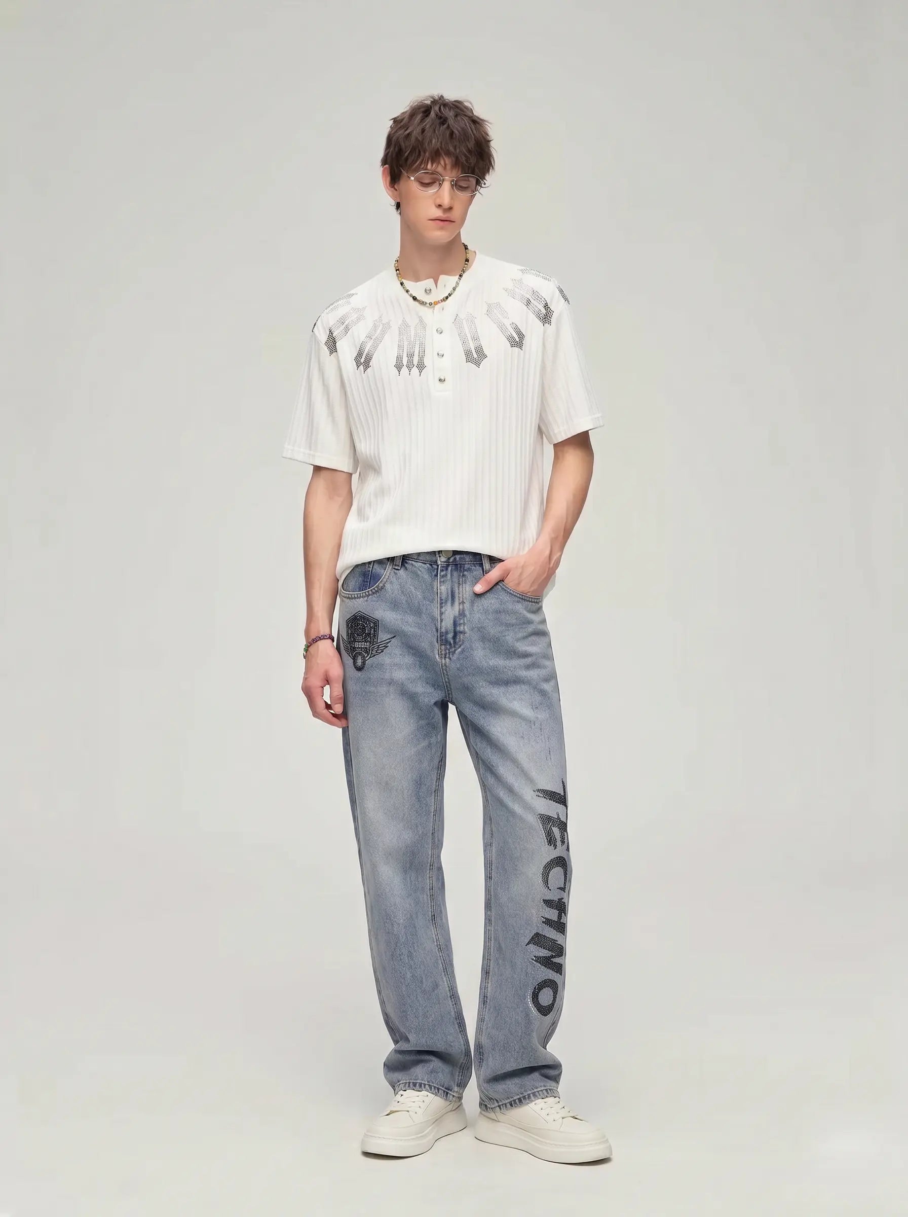 A model stands against a light gray studio background wearing a white ribbed short-sleeve henley shirt with black feather-like embroidery on the chest, light-wash blue jeans with black text down the leg, and white sneakers. TCH Gothic Rhinestone Letter Ribbed Henley T-Shirt