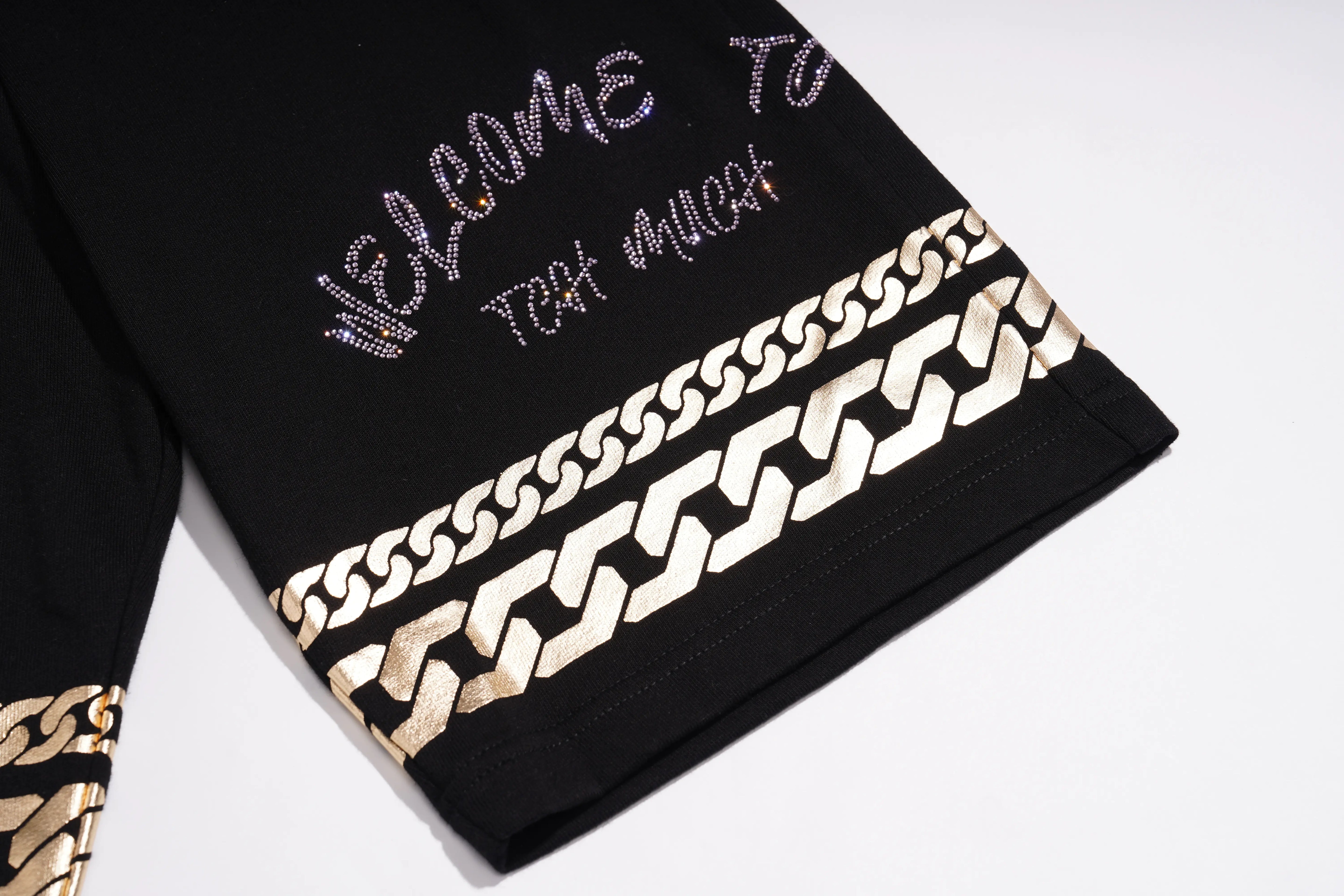 Close-up of a black garment sleeve with rhinestone-style 'Welcome to That Much' text and a wide metallic gold chain-link border, shown on a white background. TCH Gothic Rhinestone Teddy Bear Chain Print Sweat Shorts