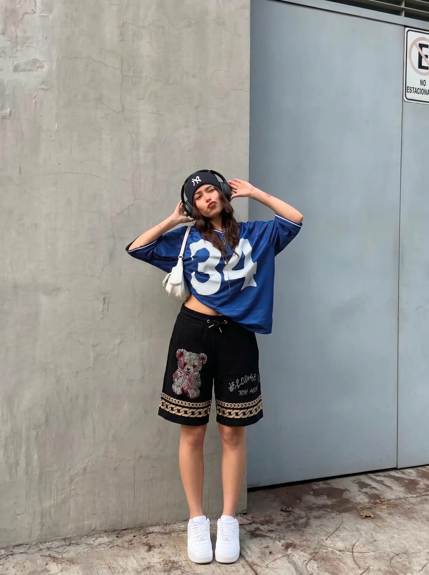 A woman posing outdoors in a blue oversized t-shirt with '34' printed on it, black shorts with a teddy bear graphic and gold trim, white sneakers, and a black beanie. TCH Gothic Rhinestone Teddy Bear Chain Print Sweat Shorts
