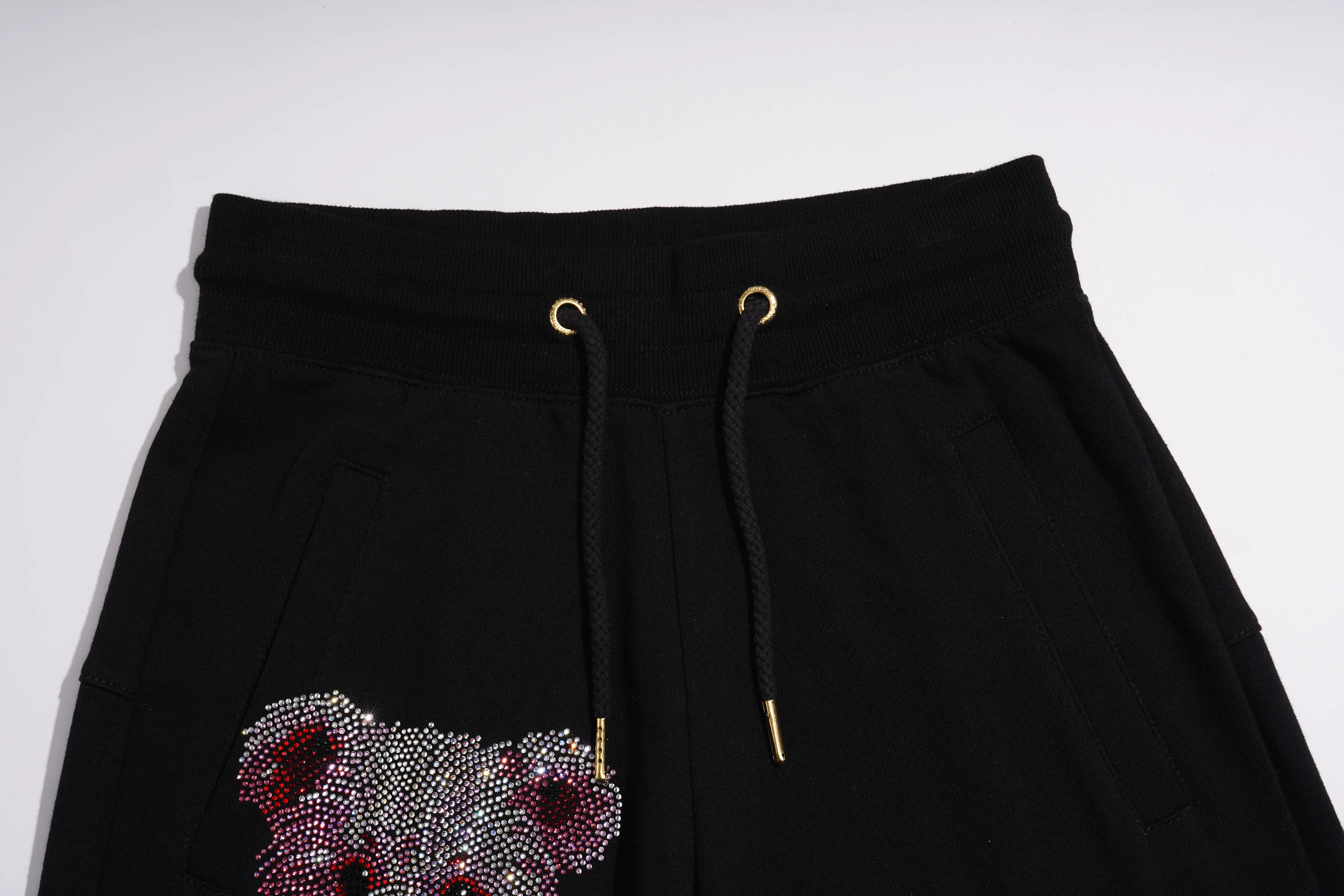 Black sweatpants featuring a prominent teddy bear face embellished with sparkling rhinestones in red, pink, and silver on the lower left leg, with black drawstrings and gold eyelets at the elastic waistband. TCH Gothic Rhinestone Teddy Bear Chain Print Sweat Shorts