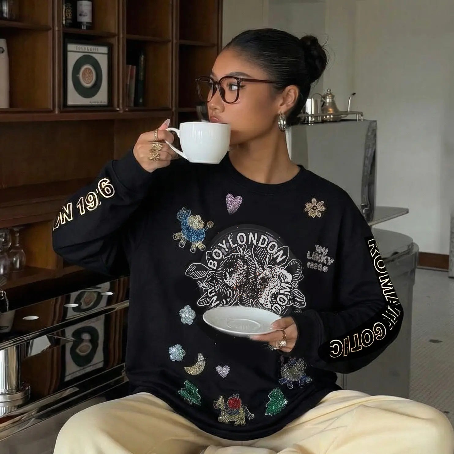 A woman wearing a black sweatshirt with colorful designs, sitting in a room holding a cup and saucer. TCH Graphic Rhinestone Heat Transfer Crewneck Sweatshirt-1
