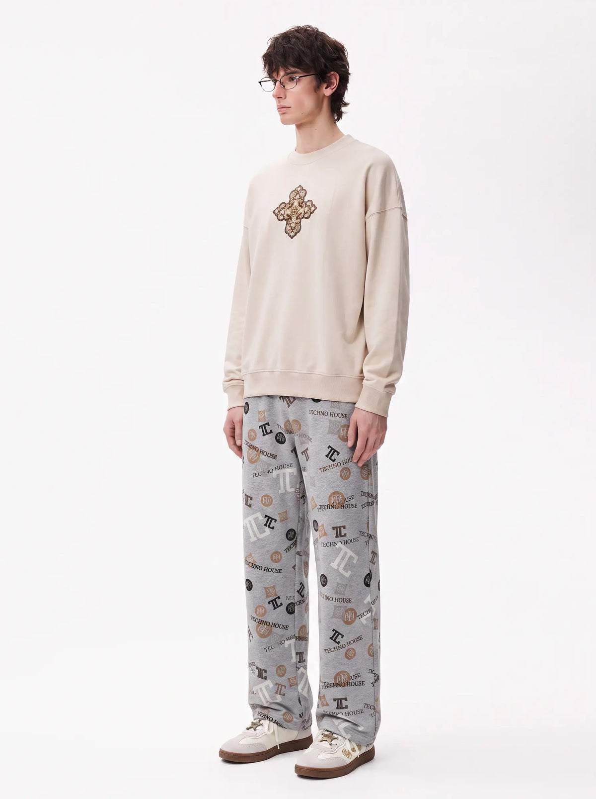 A man wearing a beige crewneck sweatshirt with a gold-toned embroidered cross on the chest, paired with gray patterned pants and white sneakers, standing against a plain white studio background. TCH Gray Techno Graphic Wide-Leg Sweatpants
