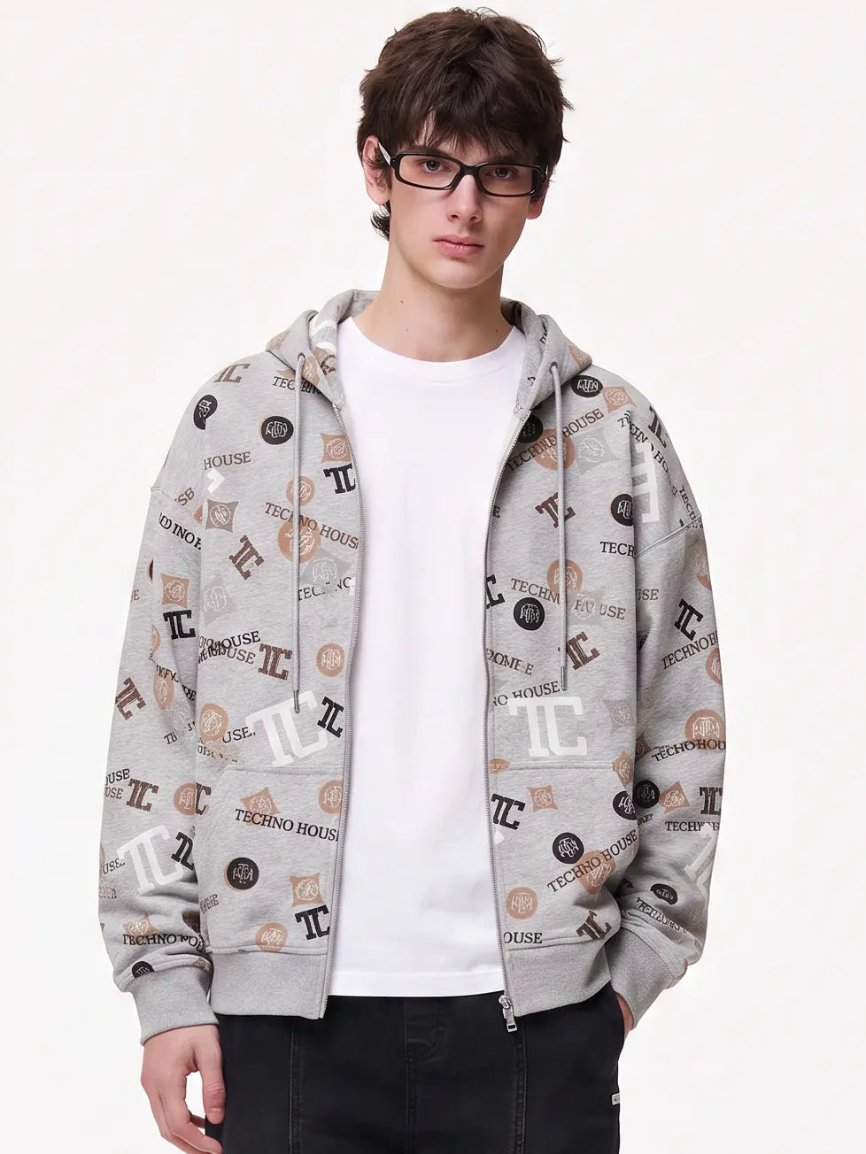 A young man wearing glasses, a light blue and white striped button-down shirt, a blue patterned necktie, and gray jeans, standing against a plain off-white studio background. TCH Grey Logo Print Zip Hoodie