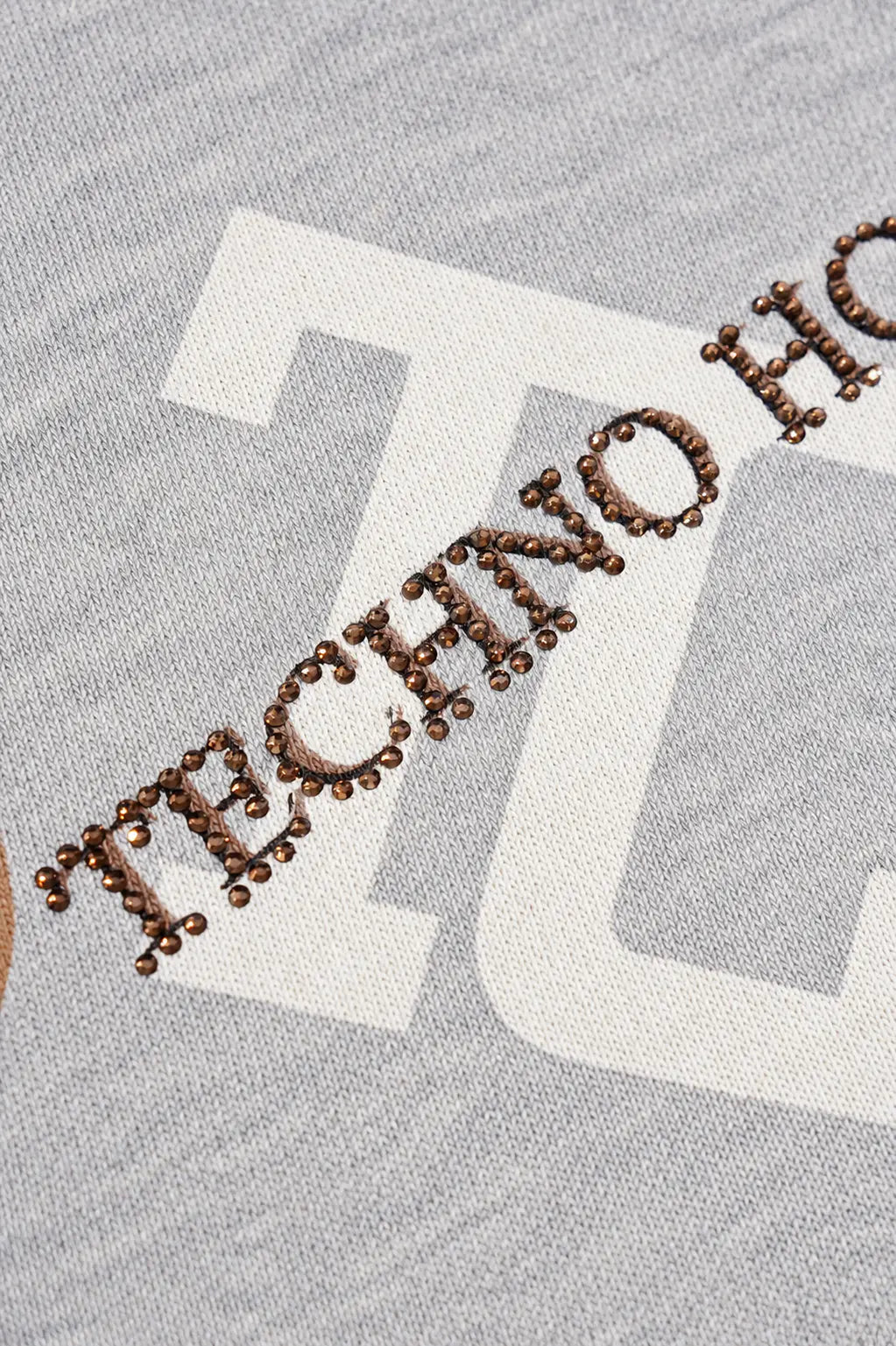 Close-up of a heather gray fabric garment with a large white graphic letter and brown embroidered text reading "TECHNO H. TCH Grey Logo Print Zip Hoodie