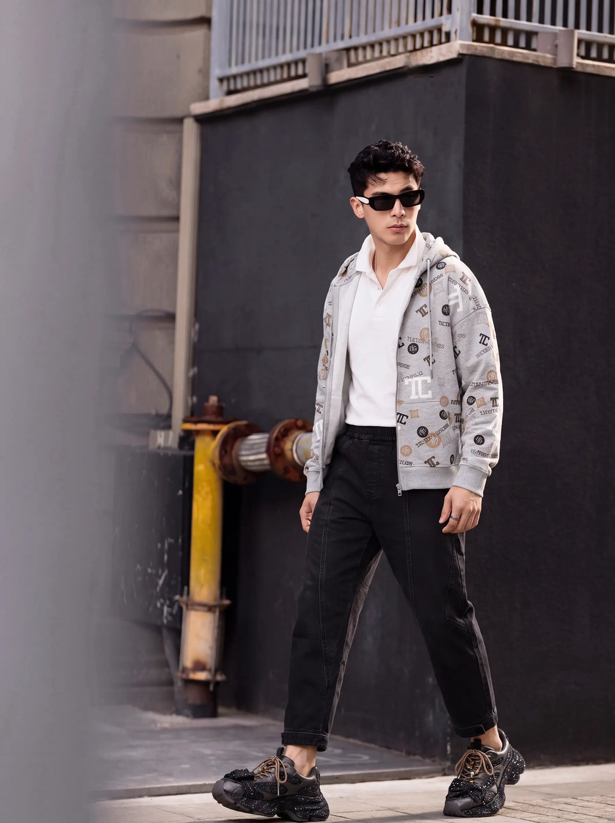 A man wearing a light gray graphic zip-up hoodie over a white polo shirt, black pants, and black sneakers, walking outdoors against a dark wall. TCH Grey Logo Print Zip Hoodie