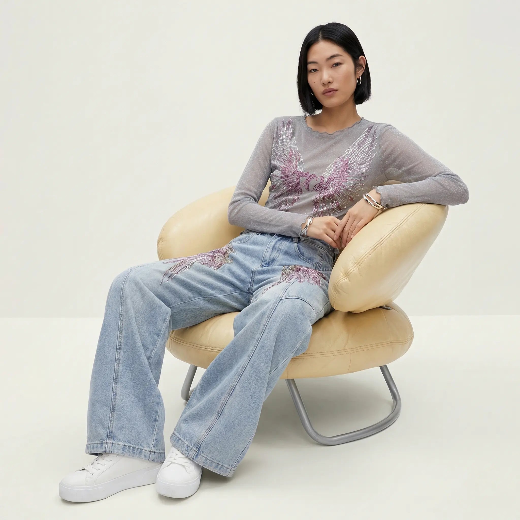 Person sitting on a modern chair wearing a gray top and light blue jeans with floral patterns. TCH Grey Rhinestone Purple Wings Long Sleeve Top
