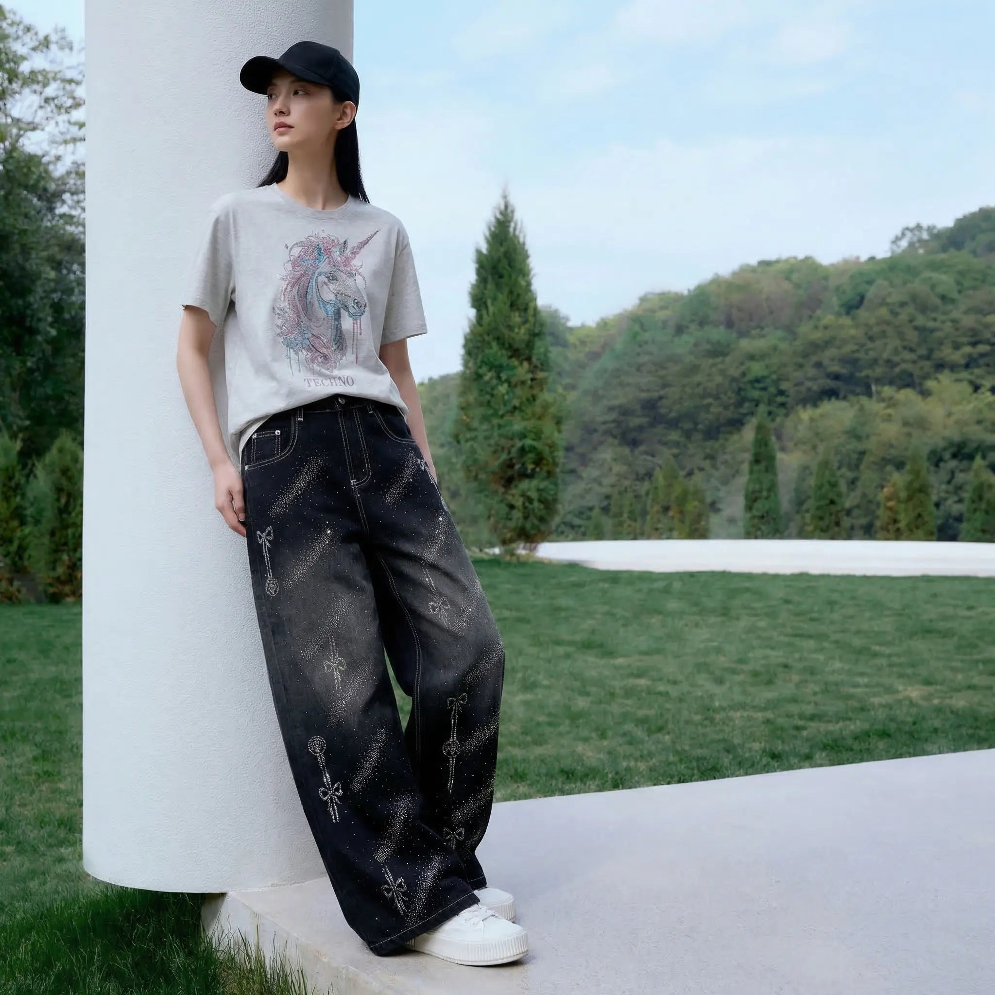 Person wearing a graphic t-shirt and black jeans standing outdoors with greenery in the background, TCH Grey Rhinestone Unicorn Graphic T-Shirt