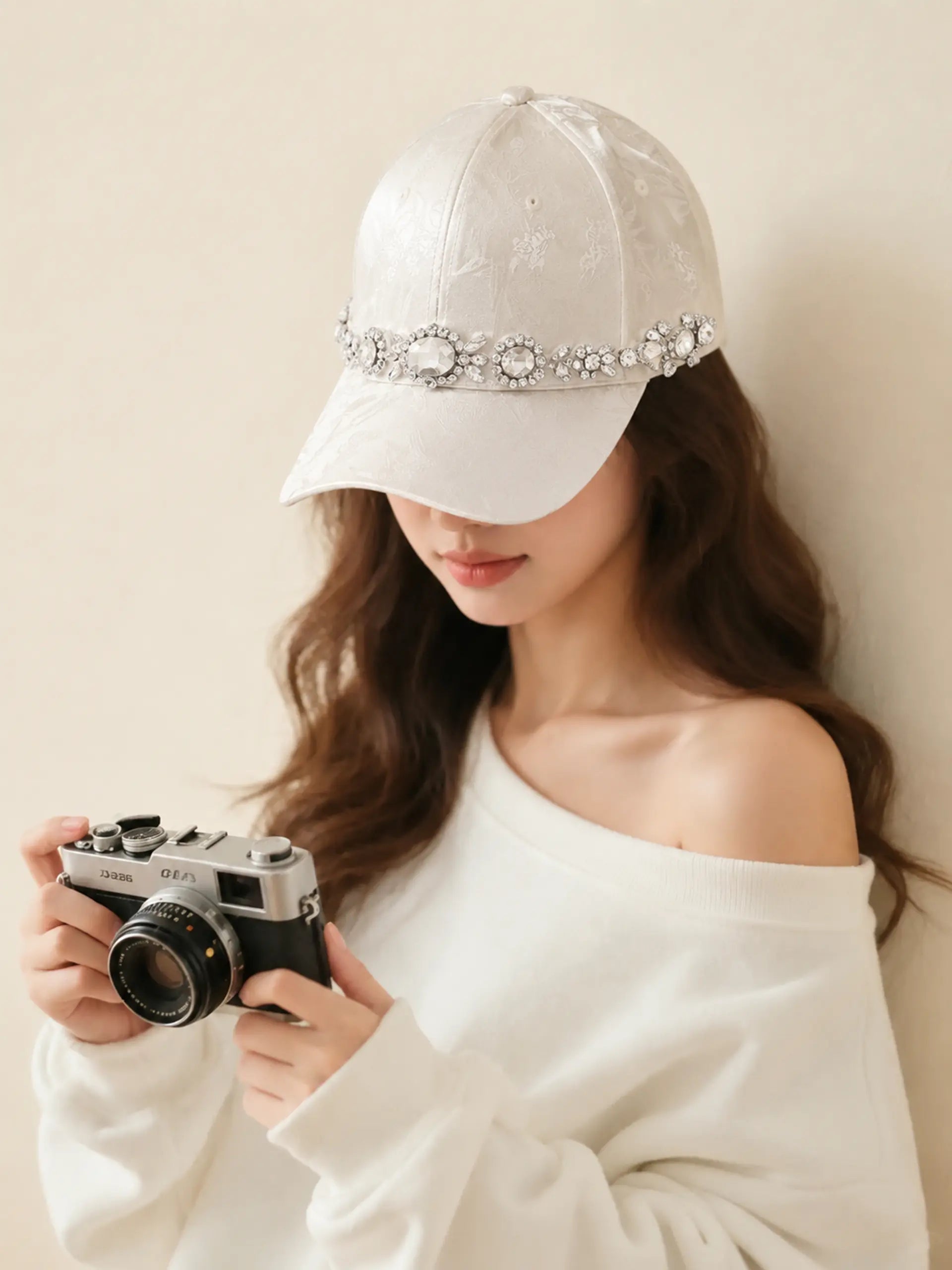 A woman wearing a white baseball cap embellished with a sparkling rhinestone band, holding a vintage silver camera against a warm beige background. TCH Luxury Crystal Embellished Floral Jacquard Baseball Cap