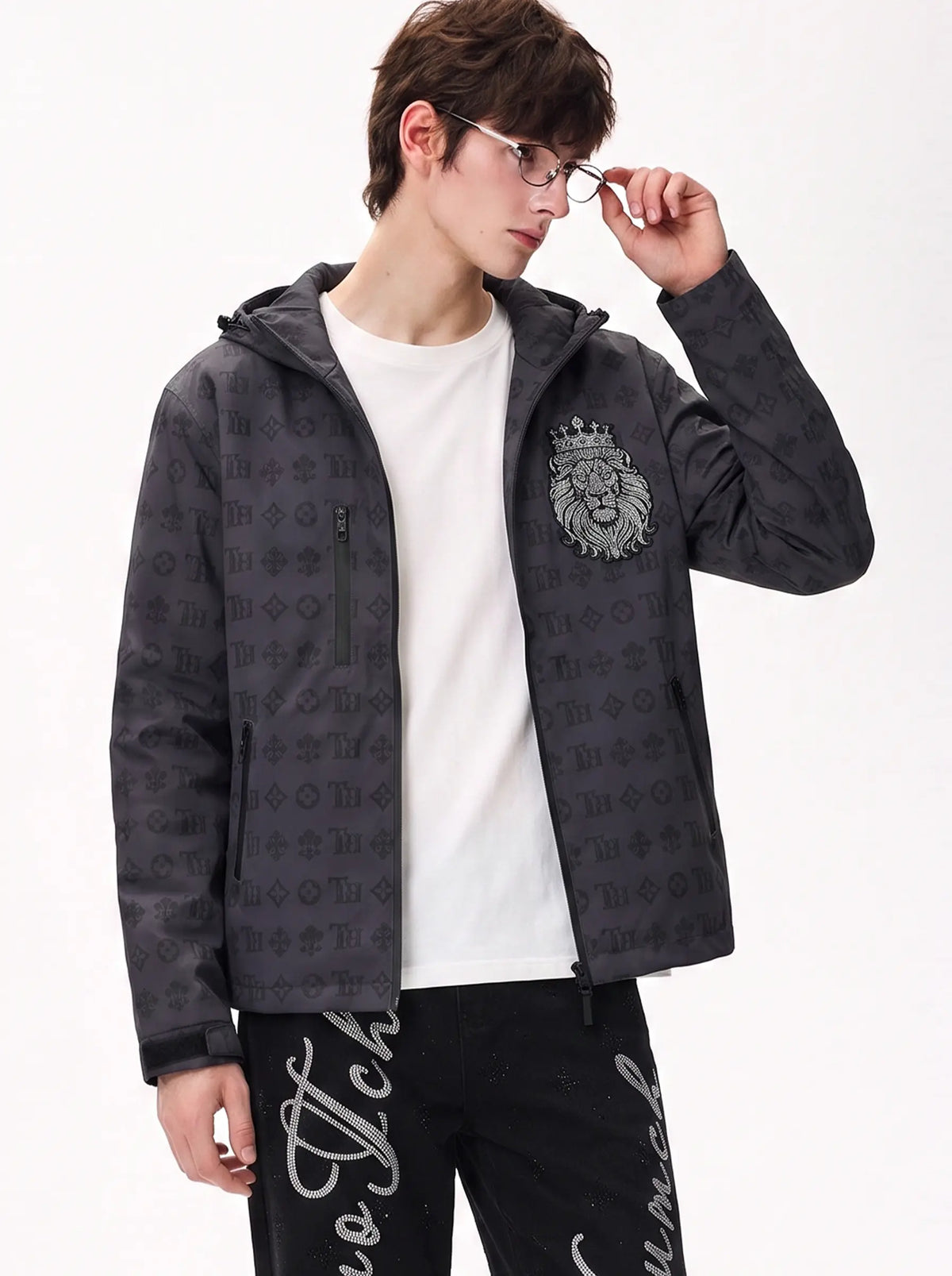 A man wearing a dark gray hooded jacket with an allover tonal monogram pattern and a large lion crest on the left chest, layered over a white t-shirt and black pants. TCH Luxury Monogram Hooded Windbreaker Jacket