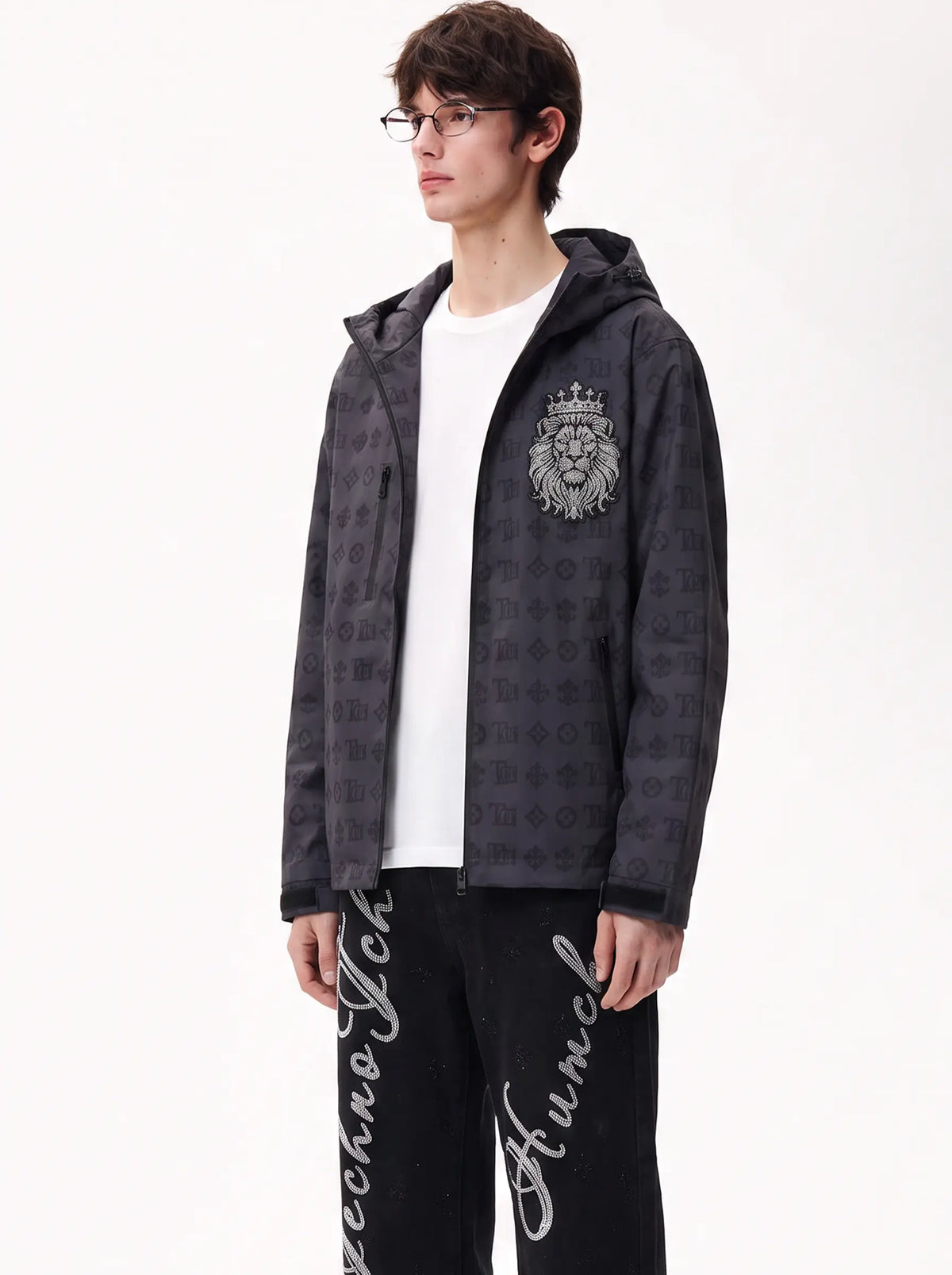 A man wearing a dark gray Louis Vuitton zip-up hoodie with an embossed monogram pattern and a silver lion crest on the chest, styled over a white t-shirt and black sweatpants with white script. TCH Luxury Monogram Hooded Windbreaker Jacket