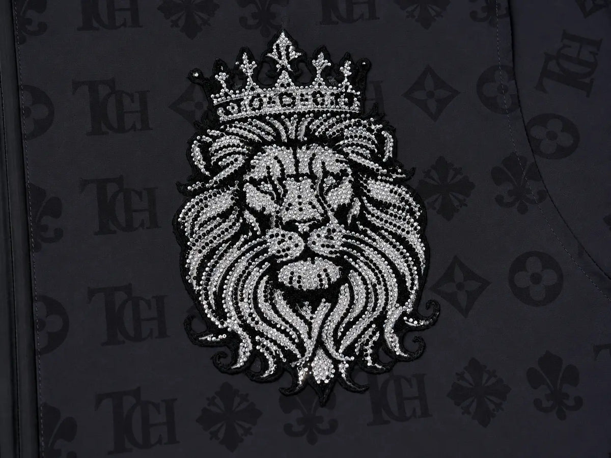 A close-up of a black garment featuring a large embroidered lion head with a crown on the chest. The embroidery is detailed with silver and white rhinestones, set against a black fabric background with a repeating monogram pattern. TCH Luxury Monogram Hooded Windbreaker Jacket