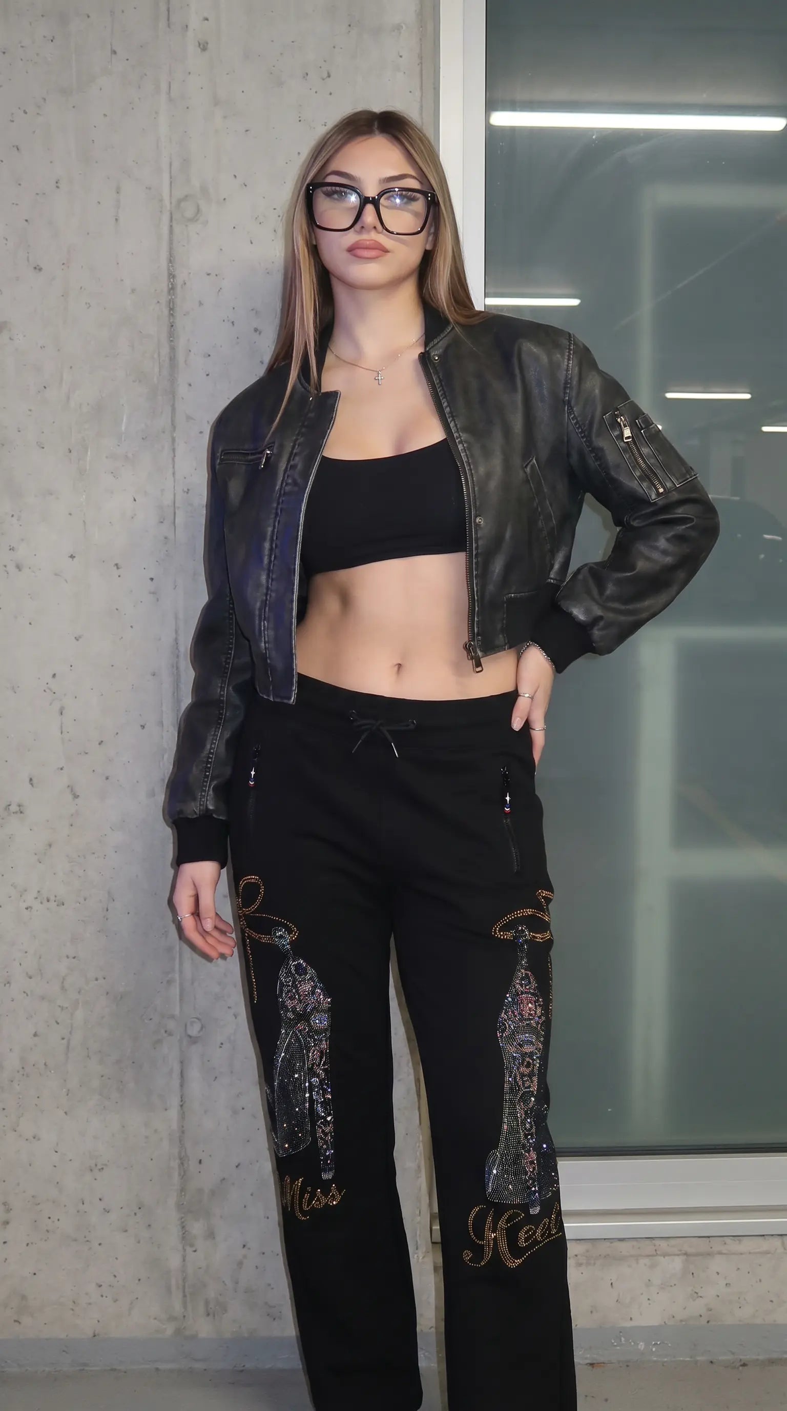 A woman modeling a black leather jacket, black crop top, and black embroidered pants with gold lettering, standing against a concrete wall. TCH Luxury Rhinestone High Heel Graphic Black Sweatpants