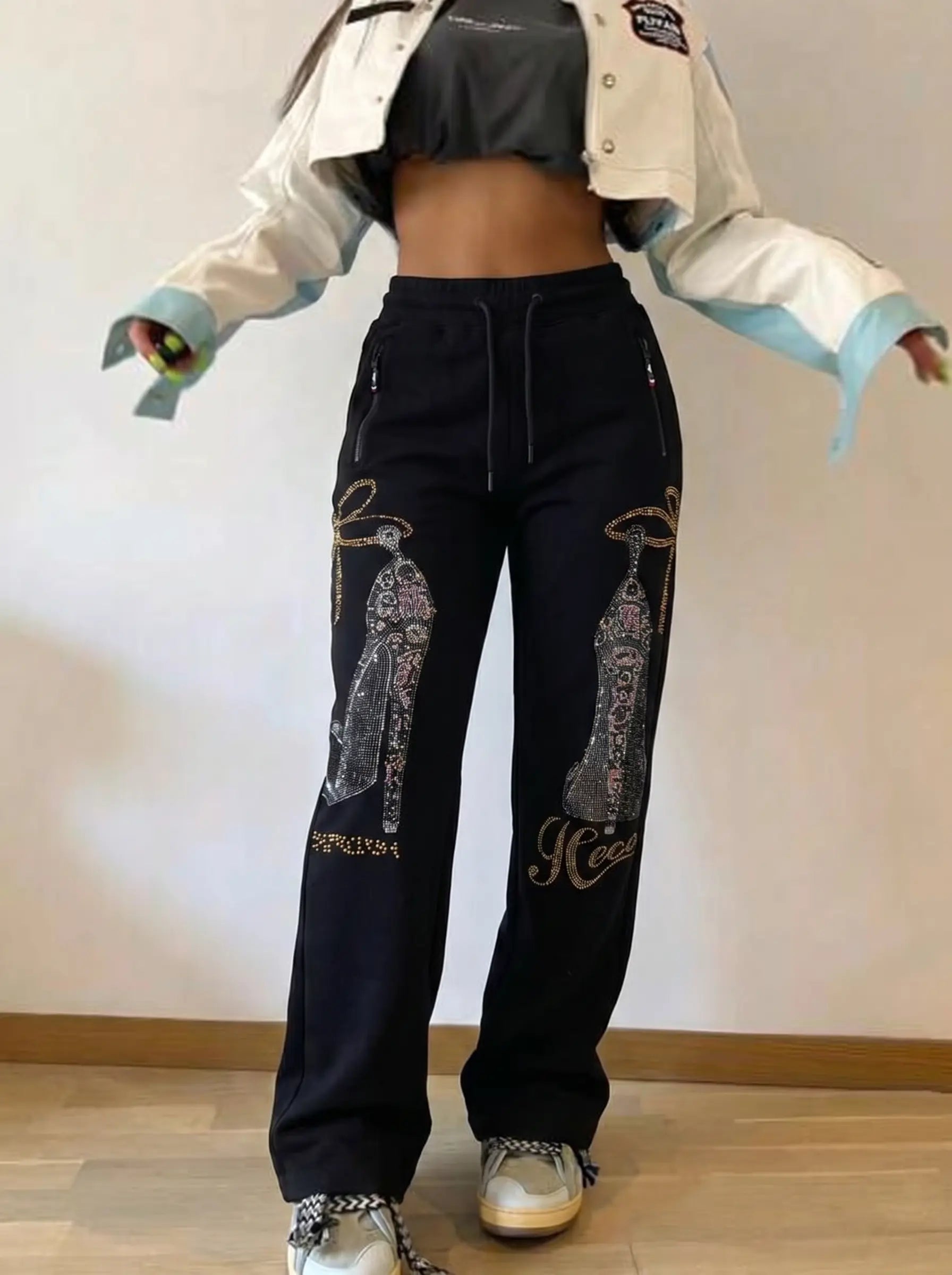 A person modeling black wide-leg sweatpants with large silver rhinestone high-heel graphics and gold script lettering on the legs, paired with a cropped white jacket, dark crop top, and sneakers, standing against a plain indoor wall. TCH Luxury Rhinestone High Heel Graphic Black Sweatpants