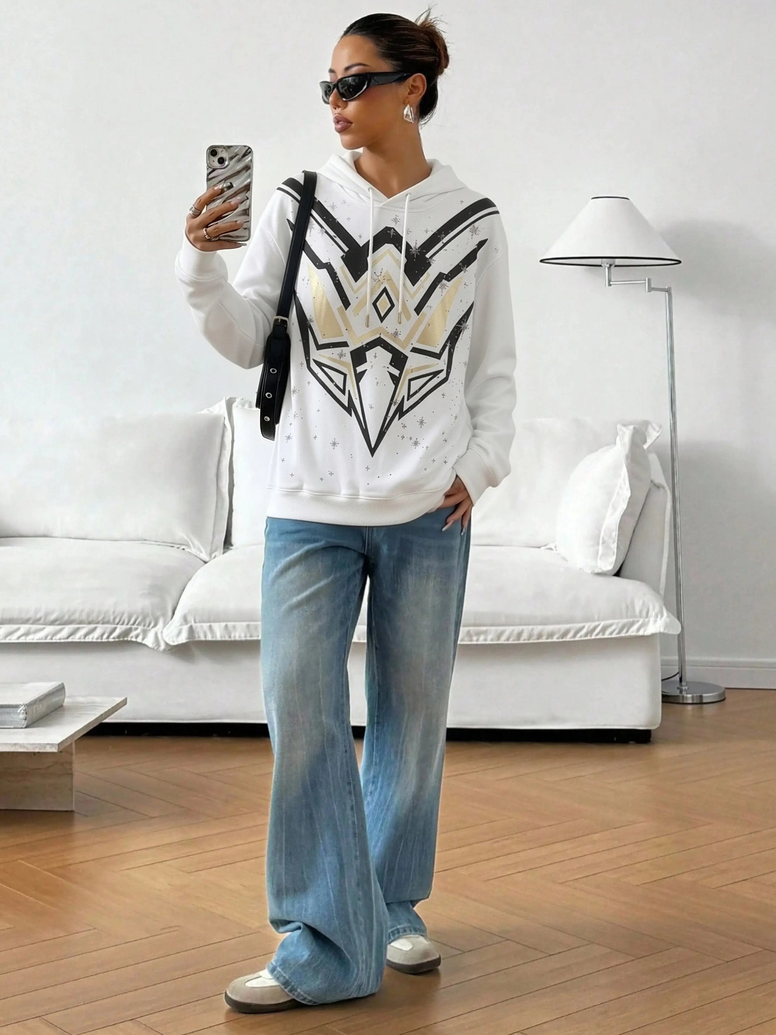 Person wearing a white hoodie with a black and gold design, blue jeans, and white sneakers in a living room. TCH Mech Graphic Rhinestone Crew Neck Sweatshirt