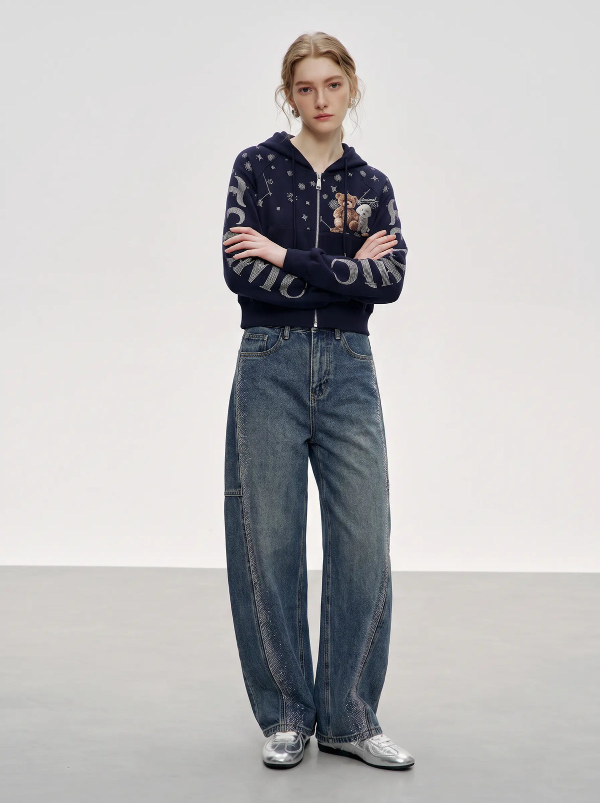 A model stands with arms crossed wearing a navy blue zip-up hoodie with white graphic lettering and a teddy bear patch, paired with wide-leg blue jeans and silver shoes, against a neutral studio background. TCH Navy Teddy Bear Rhinestone Zip Hoodie
