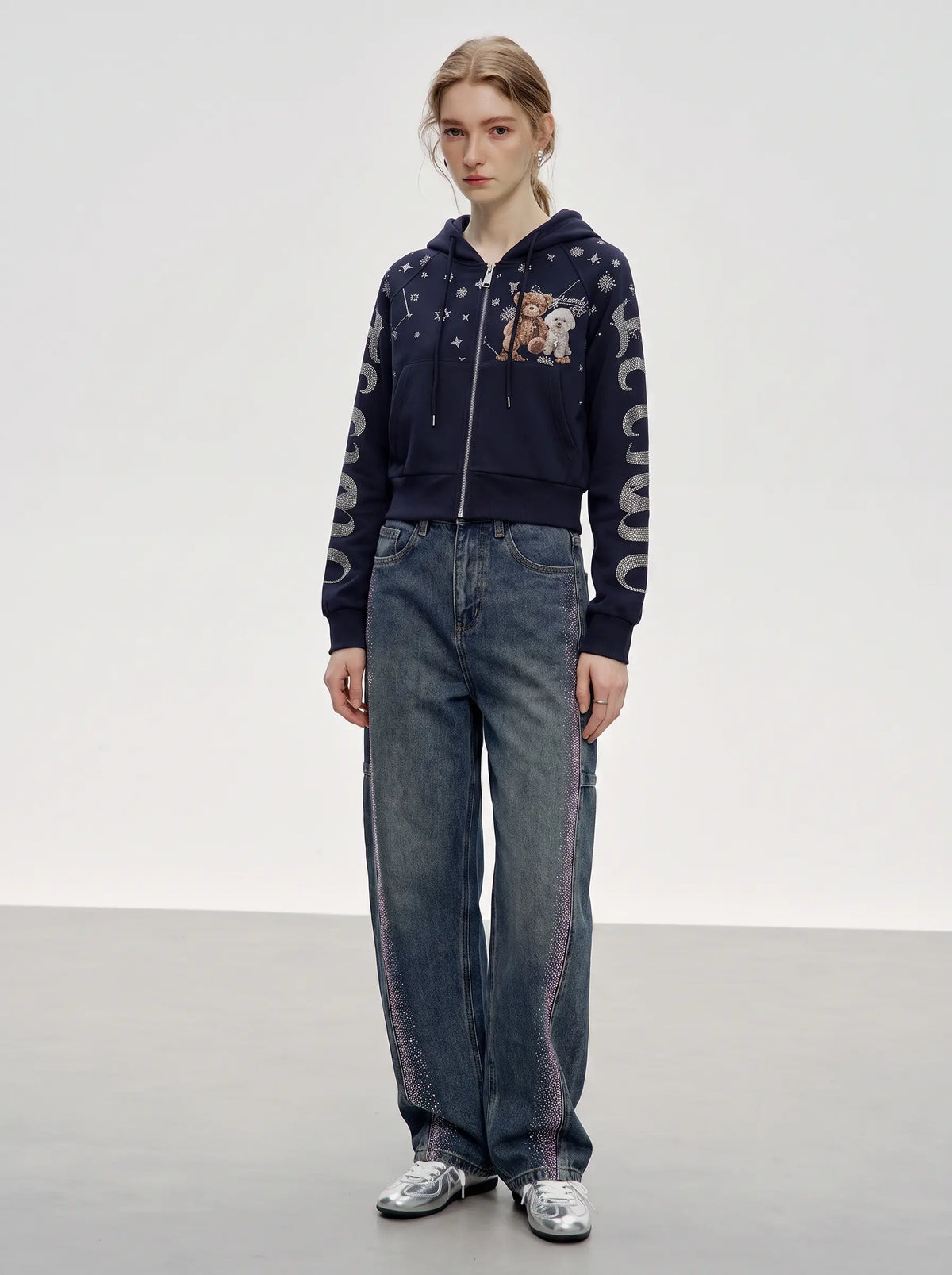 A model stands in a studio wearing a navy blue zip-up hoodie with teddy bear graphics and white lettering on the sleeves, paired with wide-leg blue jeans and silver sneakers. TCH Navy Teddy Bear Rhinestone Zip Hoodie