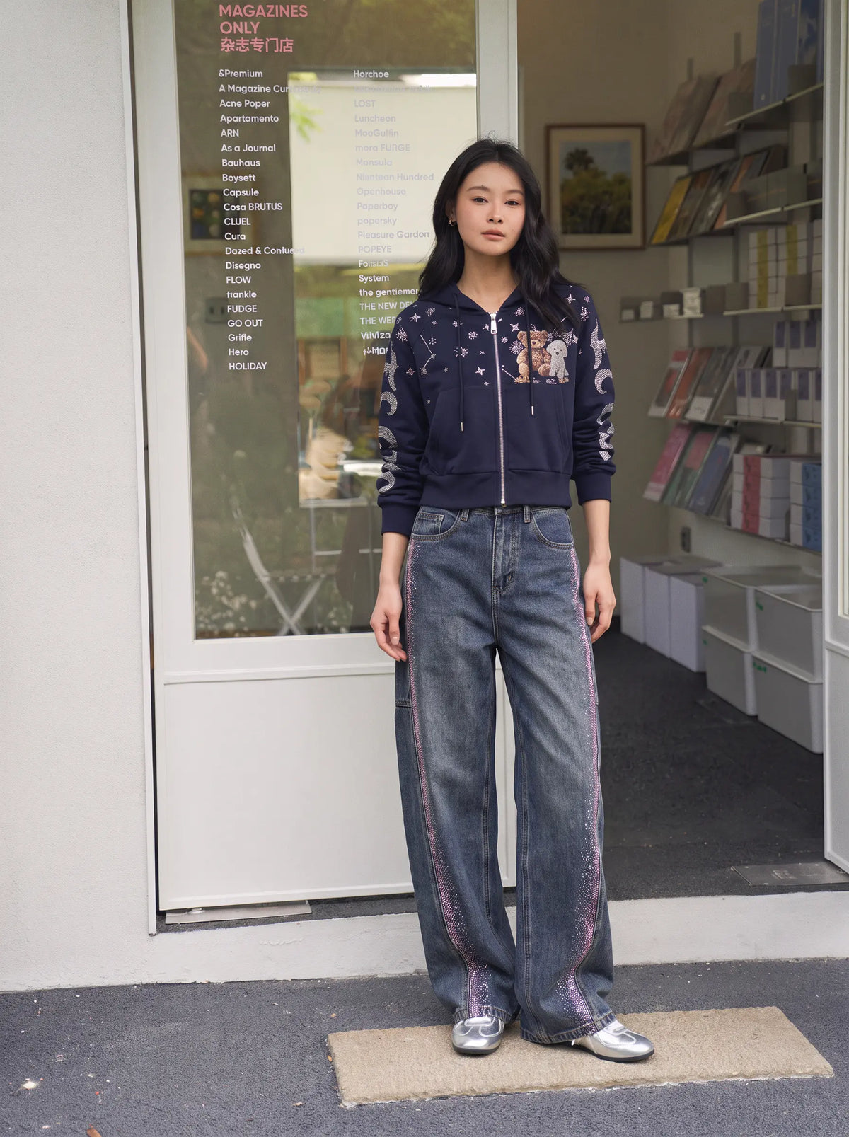 A woman stands in front of a shop doorway wearing a navy embroidered zip-up jacket, wide-leg blue jeans with pink side stripes, and silver sneakers. TCH Navy Teddy Bear Rhinestone Zip Hoodie