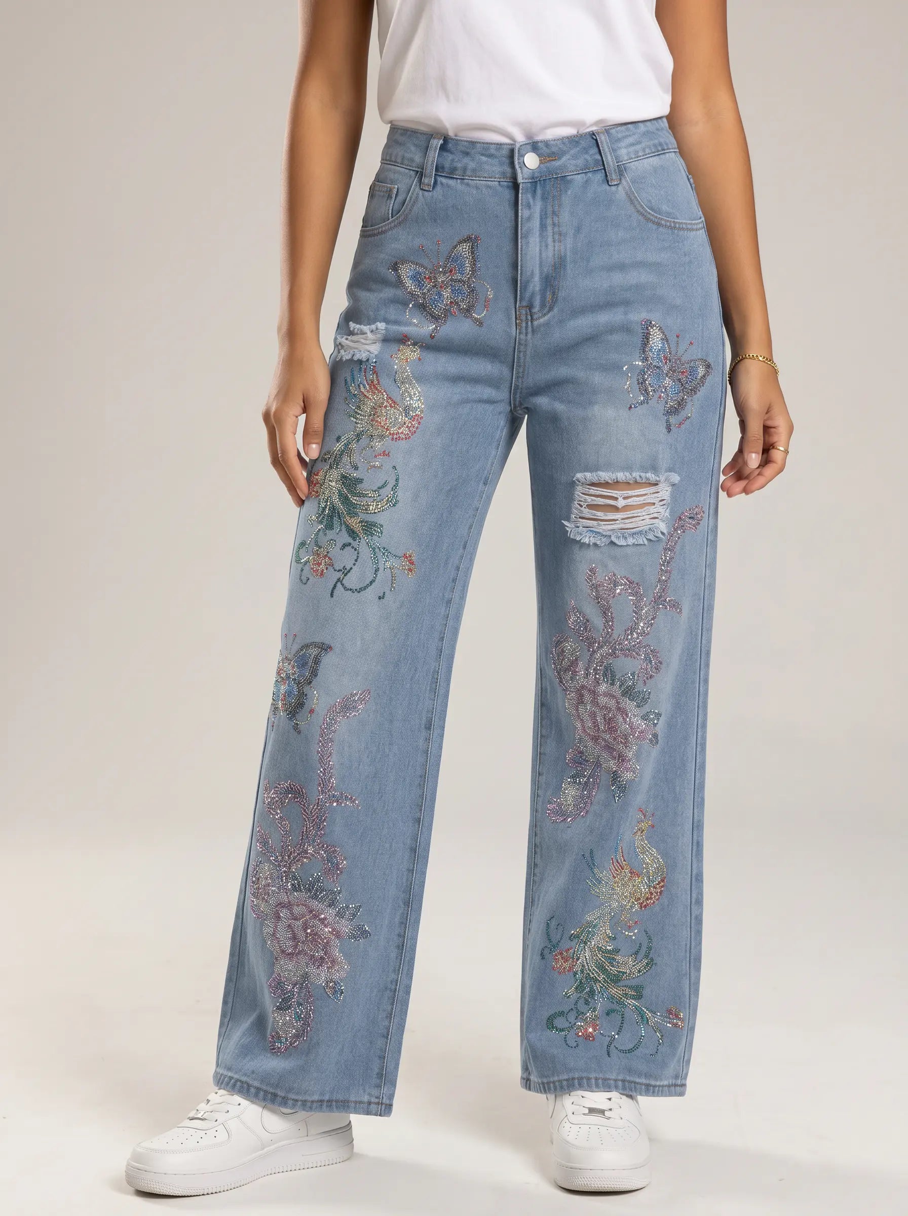 A model wearing light blue wide-leg jeans with colorful floral embroidery and distressed rips, paired with a white t-shirt and white sneakers against a neutral studio background. TCH Phoenix & Butterfly Rhinestone Wide Leg Jeans