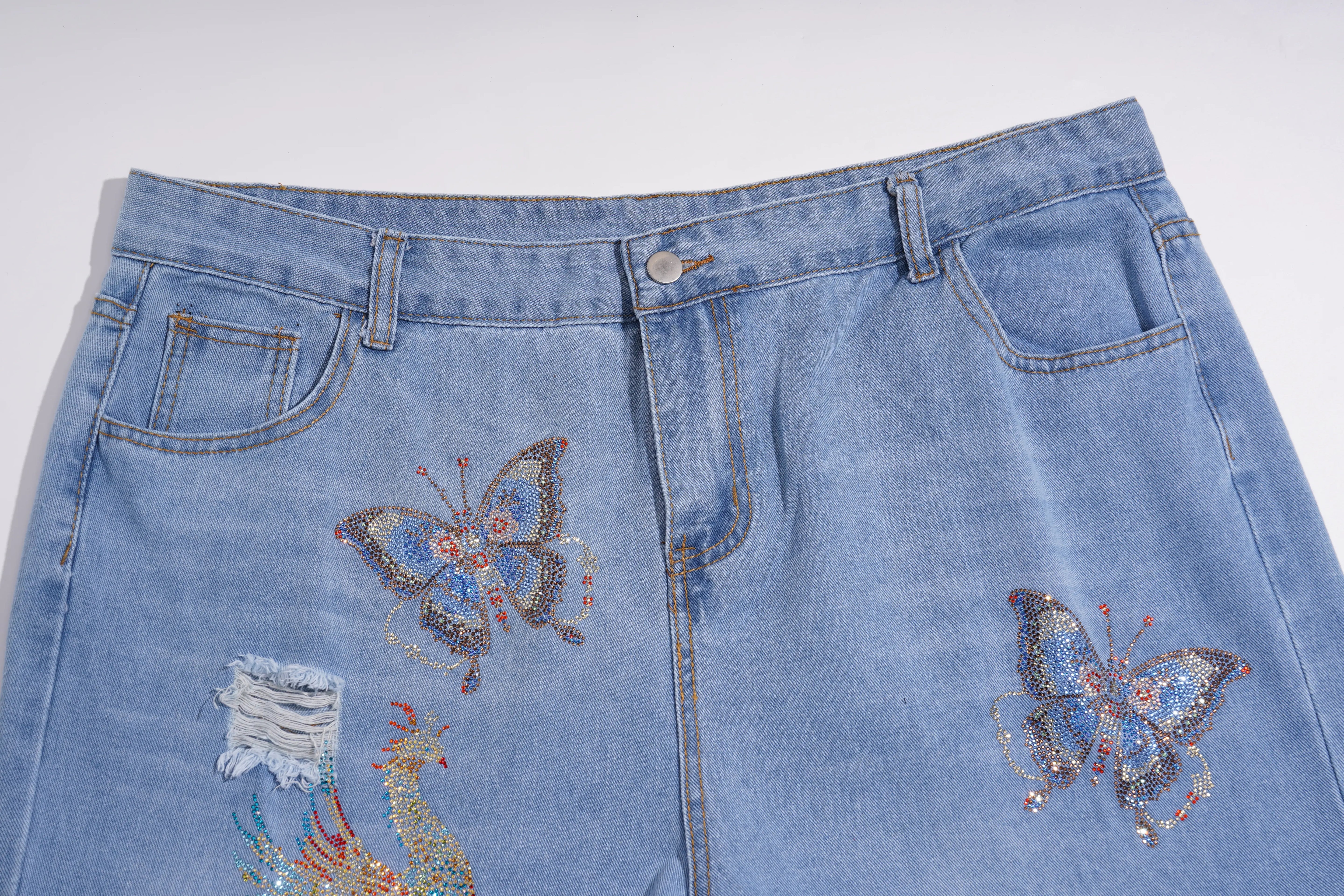 Light blue denim jeans featuring intricate butterfly and floral embroidery on the front pockets, with a distressed rip on the right leg. TCH Phoenix & Butterfly Rhinestone Wide Leg Jeans