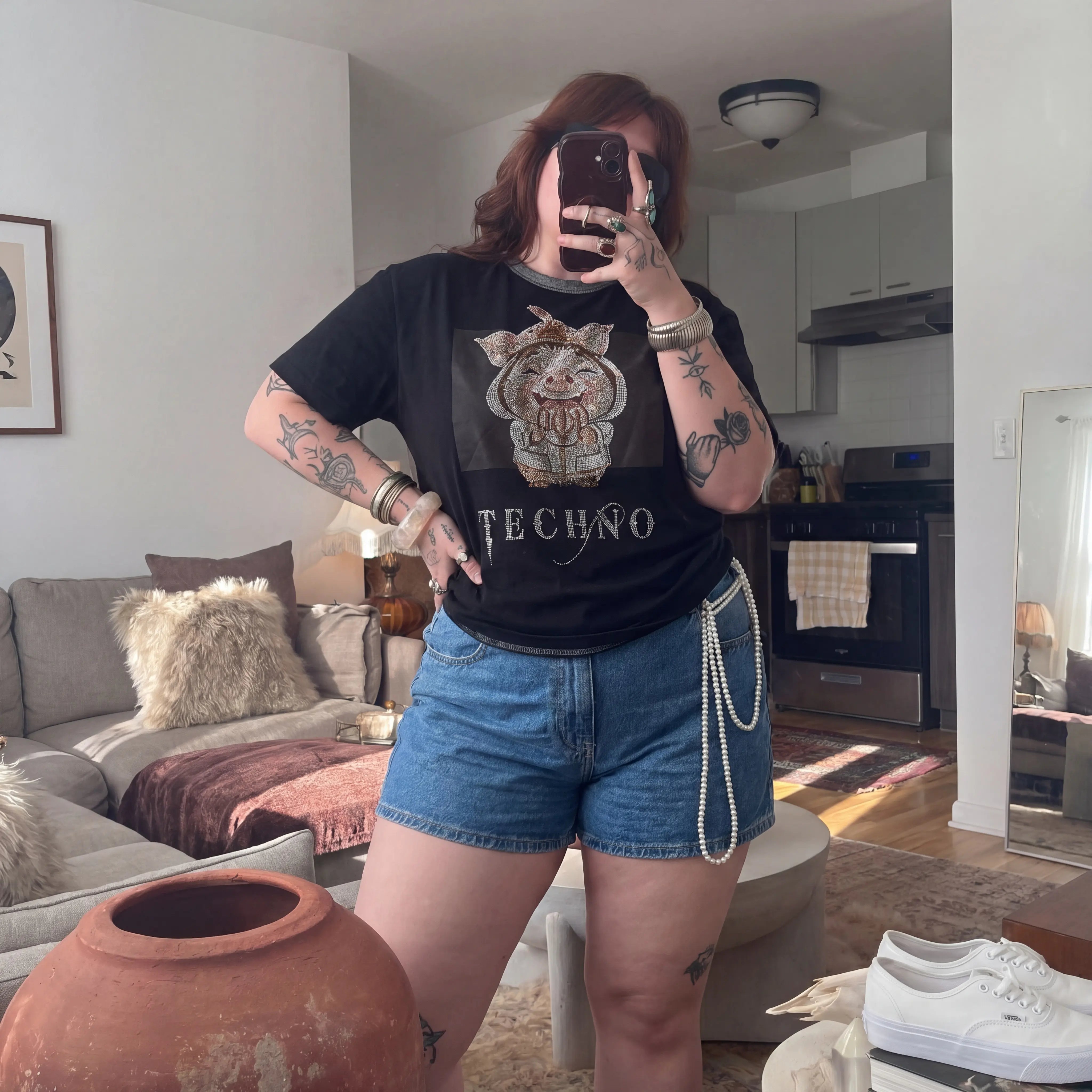 Person taking a mirror selfie in a living room wearing a black t-shirt with a graphic design and text. TCH Piggy Techno Rhinestone T-Shirt.