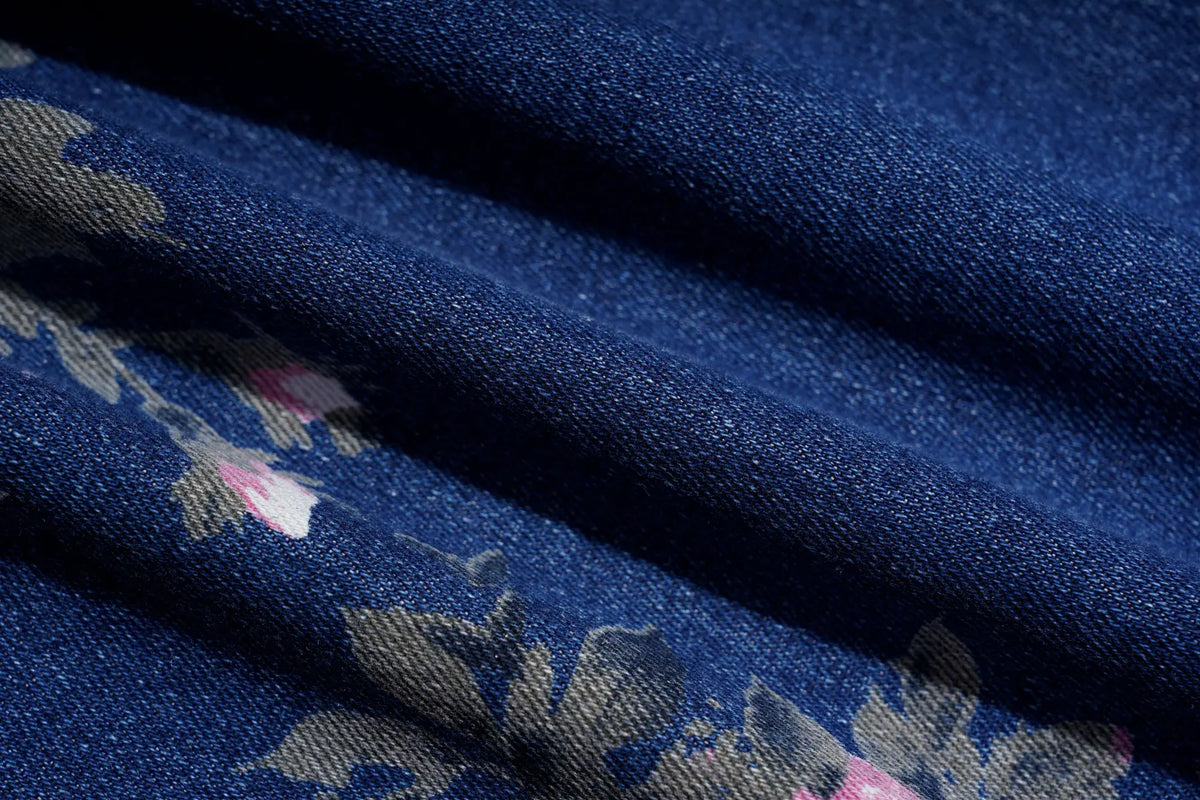 A close-up of dark blue fabric with a floral pattern featuring muted gray-green leaves and soft pink blossoms, draped in folds to show texture. TCH Pink Floral Teddy Bear Graphic Wide-Leg Jeans