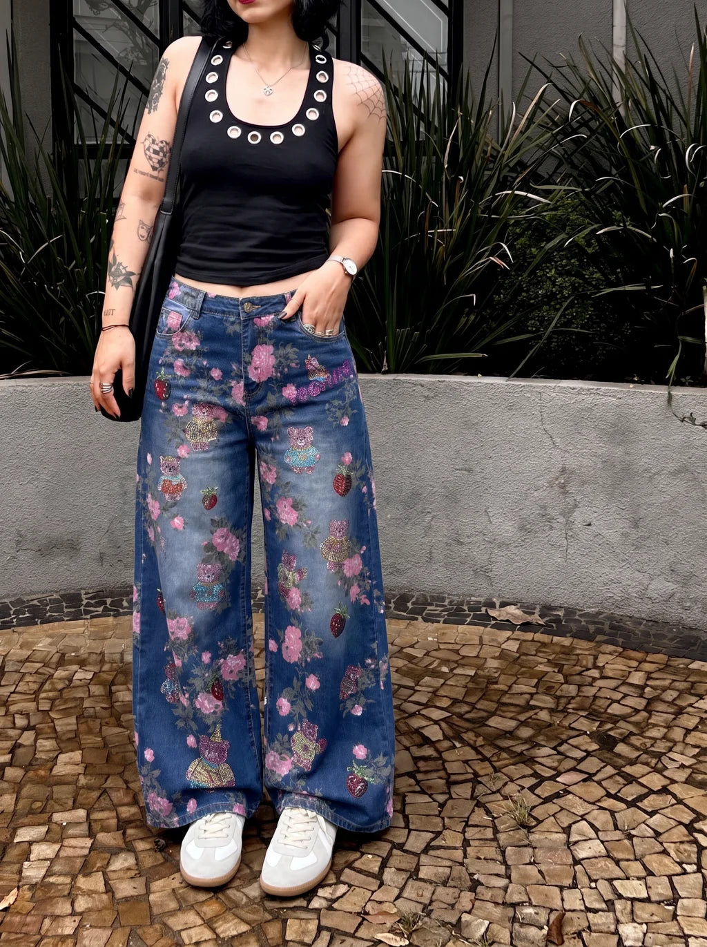 A person wearing blue wide-leg floral denim jeans, a black sleeveless top, and white sneakers, standing outdoors on a mosaic stone floor with a concrete wall and green plants behind them. TCH Pink Floral Teddy Bear Graphic Wide-Leg Jeans