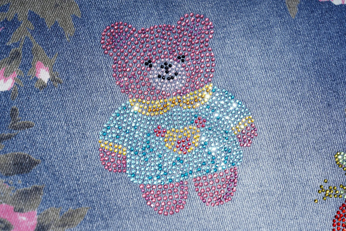 A close-up of a blue denim fabric featuring a teddy bear design made of colorful rhinestones. The bear is wearing a light blue sweater with pink and yellow accents. TCH Pink Floral Teddy Bear Graphic Wide-Leg Jeans