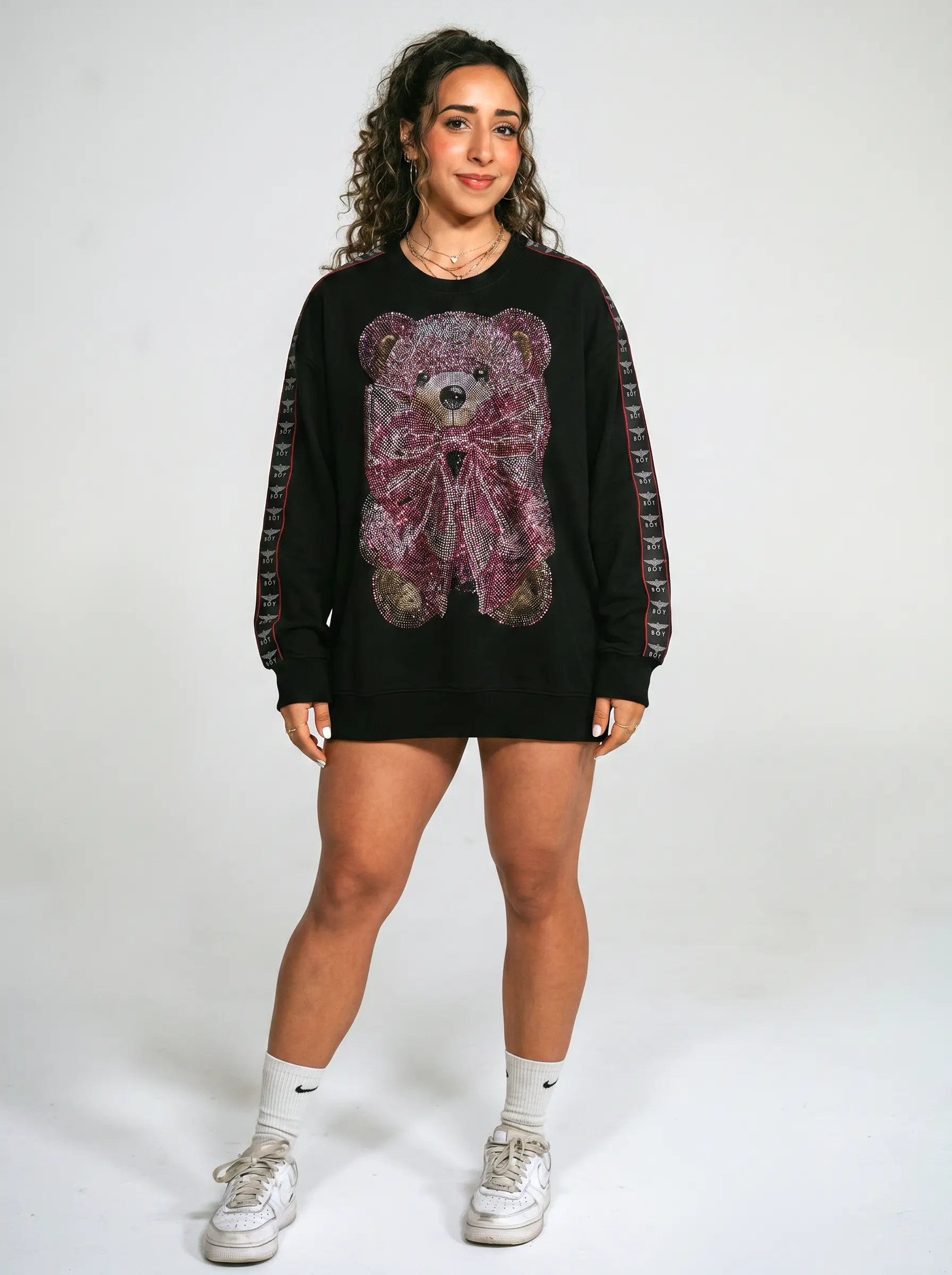 A woman modeling a black crewneck sweatshirt featuring a large teddy bear graphic on the front with pink and purple paisley details, patterned sheer sleeves, paired with white socks and sneakers against a light gray studio background. TCH Pink Teddy Bear Rhinestone Graphic Black Sweatshirt