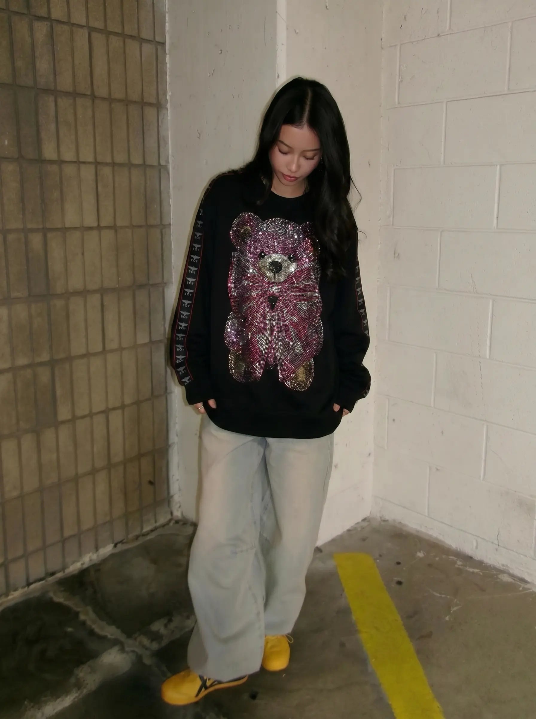A woman wearing a black crewneck sweatshirt with a large, colorful sequined teddy bear graphic on the front, paired with light gray wide-leg sweatpants and yellow sneakers, standing in a concrete parking structure. TCH Pink Teddy Bear Rhinestone Graphic Black Sweatshirt