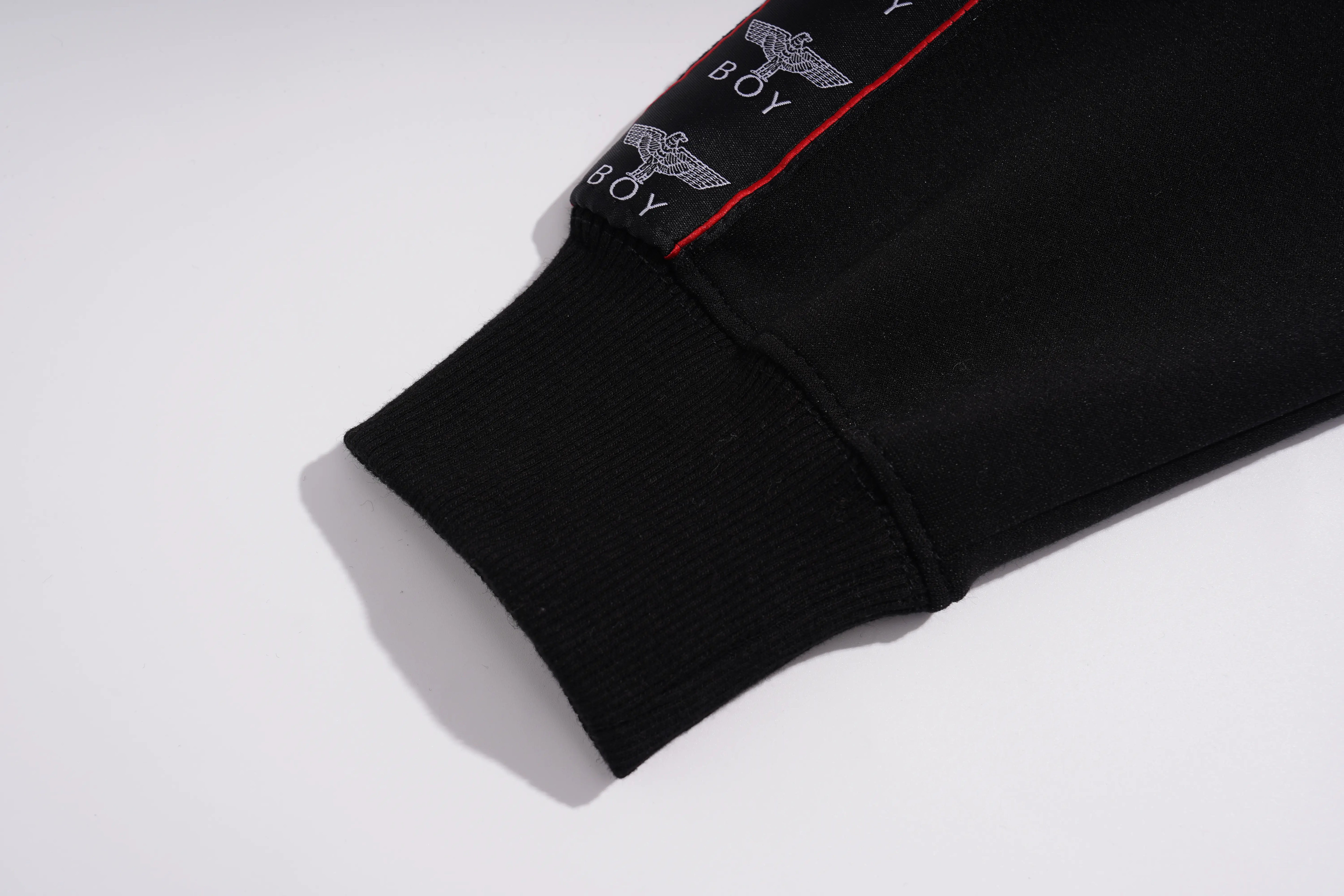 Close-up of a black ribbed sweatshirt cuff featuring a white embroidered BOY logo with an eagle graphic and a thin red accent line, on a light gray background. TCH Pink Teddy Bear Rhinestone Graphic Black Sweatshirt