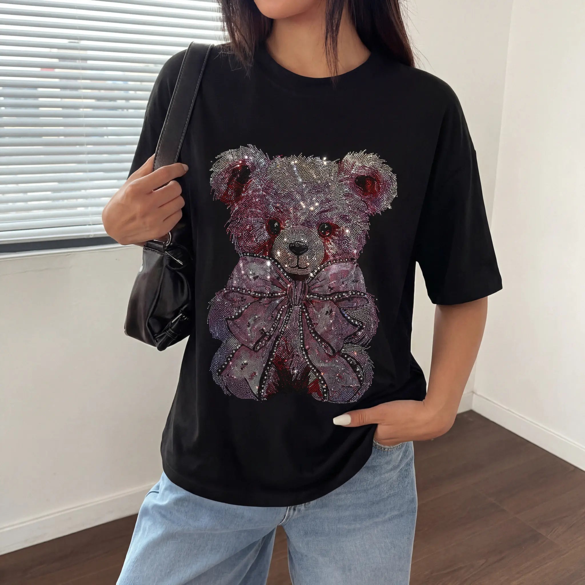 Person wearing a black t-shirt with a decorative teddy bear design, holding a black handbag. TCH Pink Teddy Bear Rhinestone T-Shirt