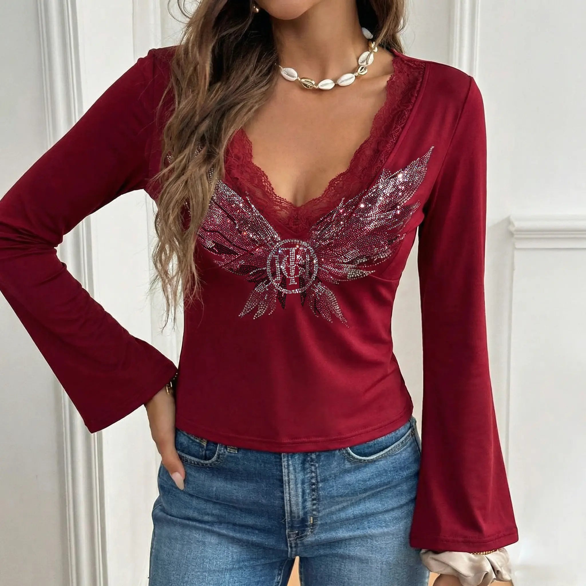 Red long-sleeve top with decorative wings and blue jeans against a white wall. TCH Red Lace Trim V-Neck Rhinestone Wing Long Sleeve Top
