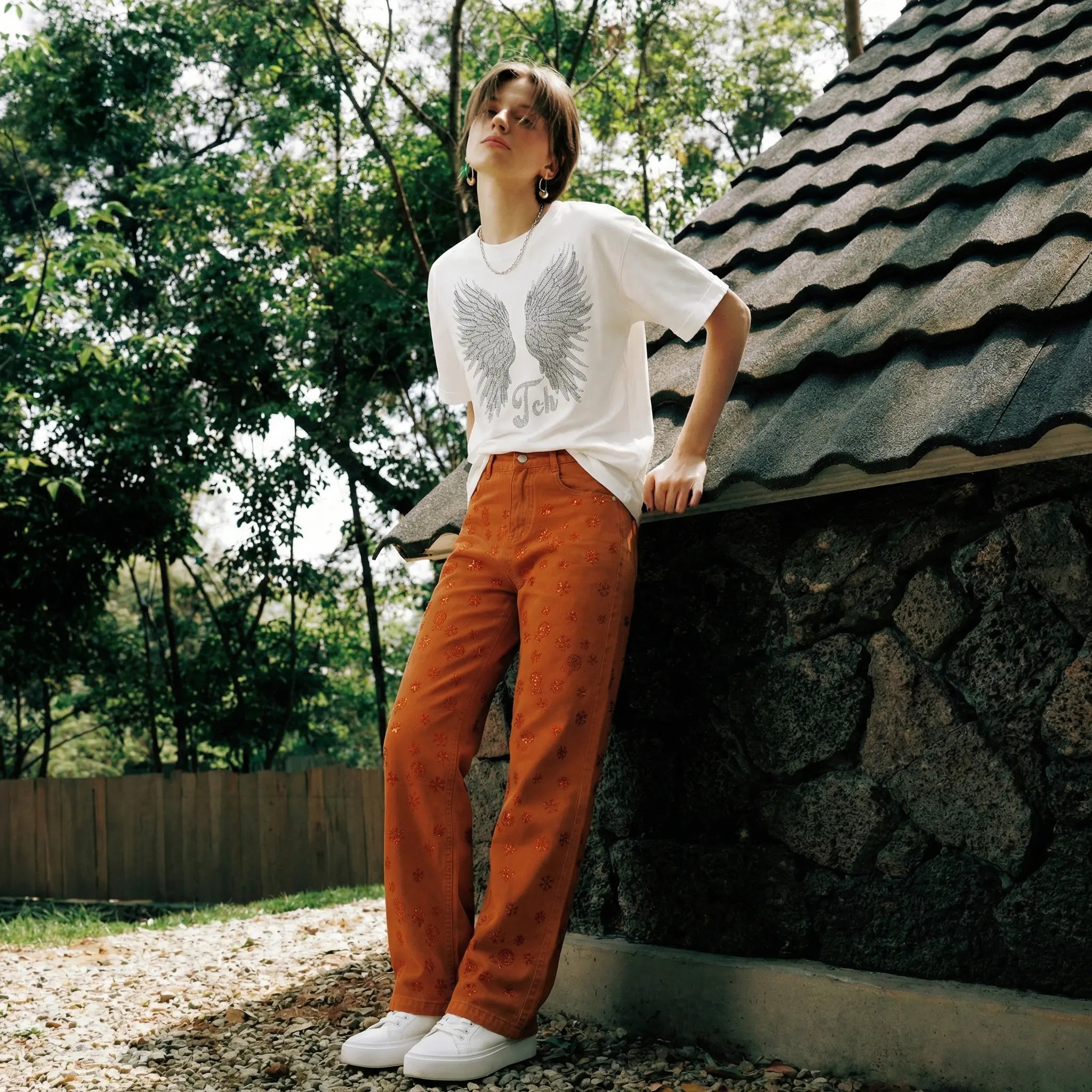 Person wearing a white t-shirt with an eagle design and orange pants standing outdoors near a stone building. TCH Red Snowflake Rhinestone Brown Denim Jeans