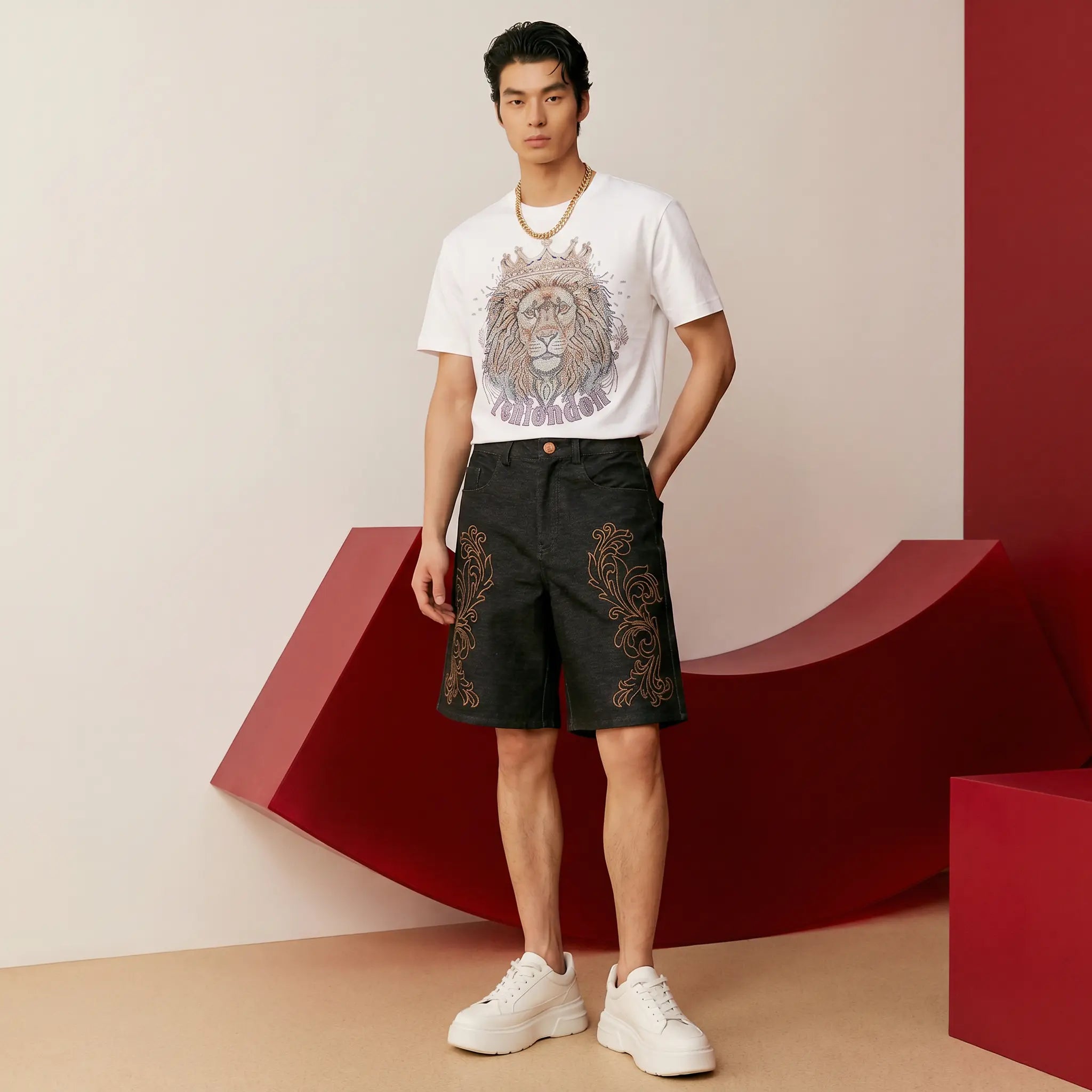 A male model stands in a studio setting wearing a white graphic T-shirt with a large lion print, black shorts with gold embroidery, and white sneakers. The background features a minimalist beige wall with bold red geometric blocks. TCH Regal Crown Lion Rhinestone Graphic T-Shirt