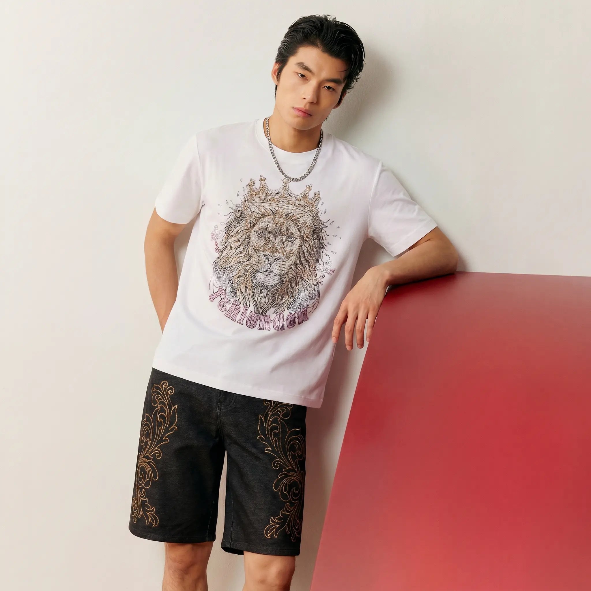 A man wearing a white graphic t-shirt with a detailed lion print and black shorts with gold patterns, leaning against a red block on a light gray wall. TCH Regal Crown Lion Rhinestone Graphic T-Shirt