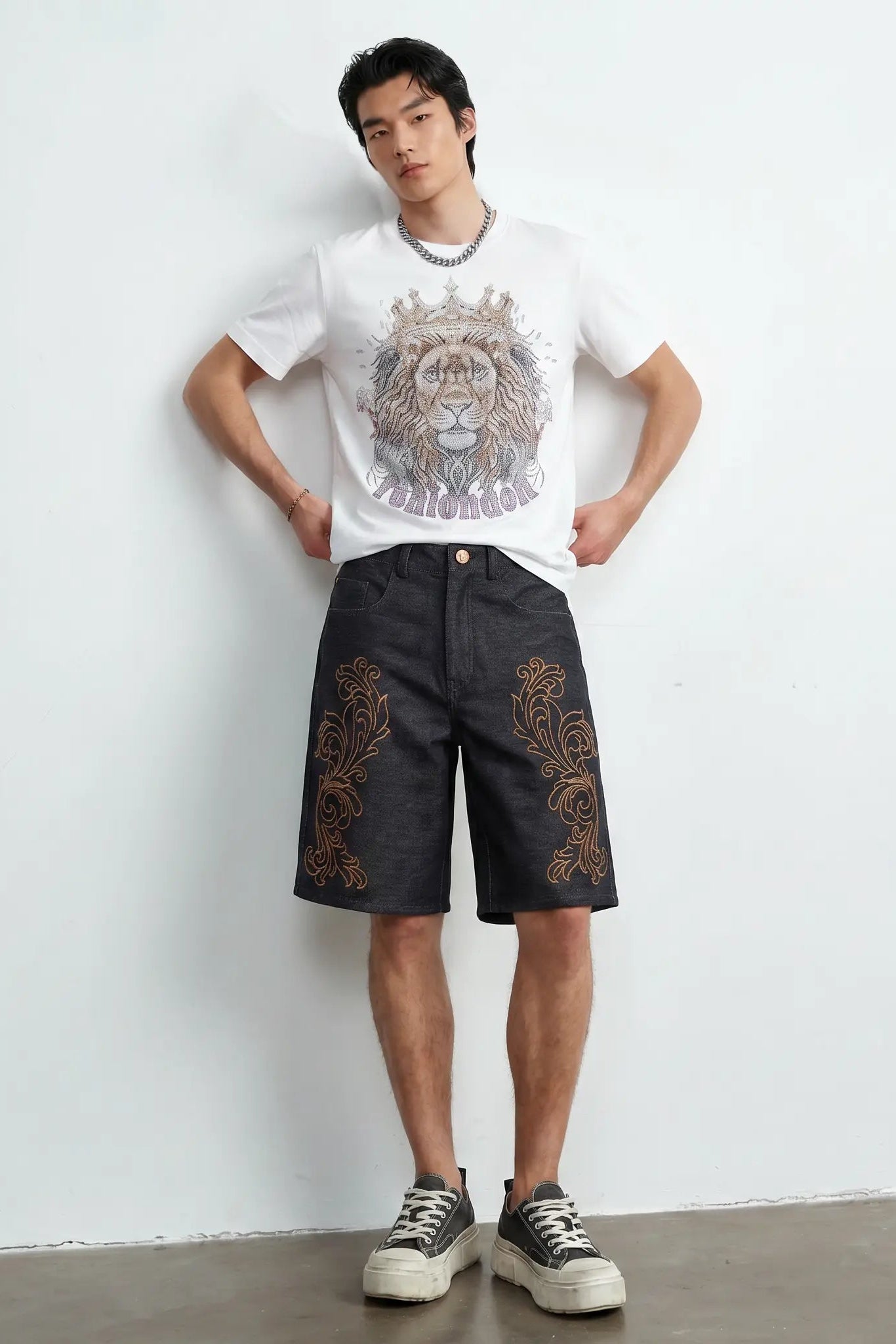 A man stands against a white wall wearing a white graphic T-shirt with a lion print, dark gray shorts with gold embroidery, and white platform sneakers. TCH Regal Crown Lion Rhinestone Graphic T-Shirt