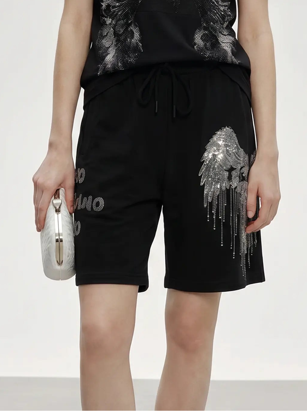 A model stands against a light gray studio background wearing a black short-sleeve T-shirt and matching black shorts, both featuring large distressed silver wing graphics. The model is holding a small metallic clutch. TCH Rhinestone Angel Wing Graphic Shorts