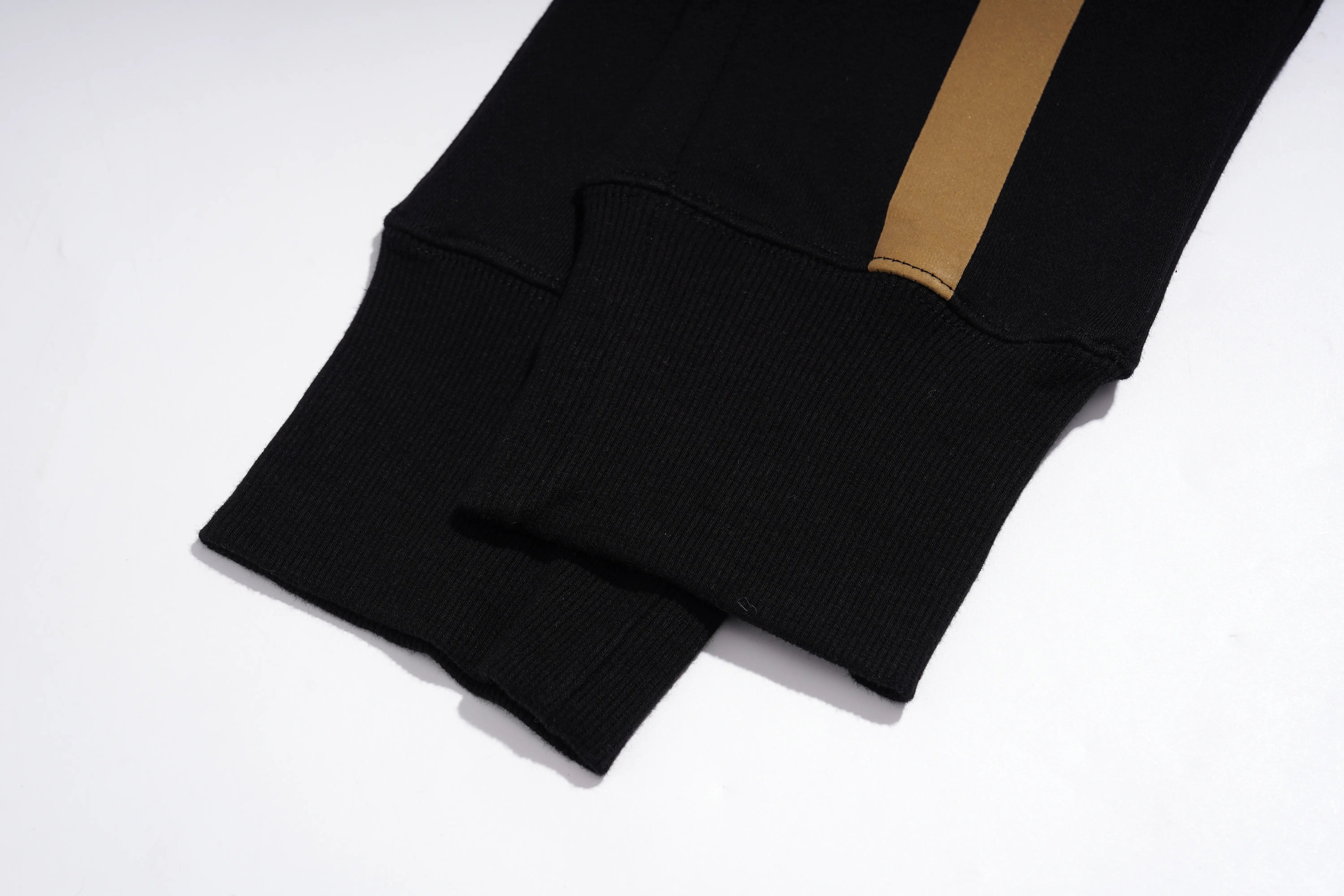 Close-up of a black ribbed knit garment, likely a sweatshirt or jacket sleeve, featuring a vertical tan or camel-colored stripe near the cuff, photographed on a white background. TCH Rhinestone Angel Wings Gold Detail Black Jogger Pants
