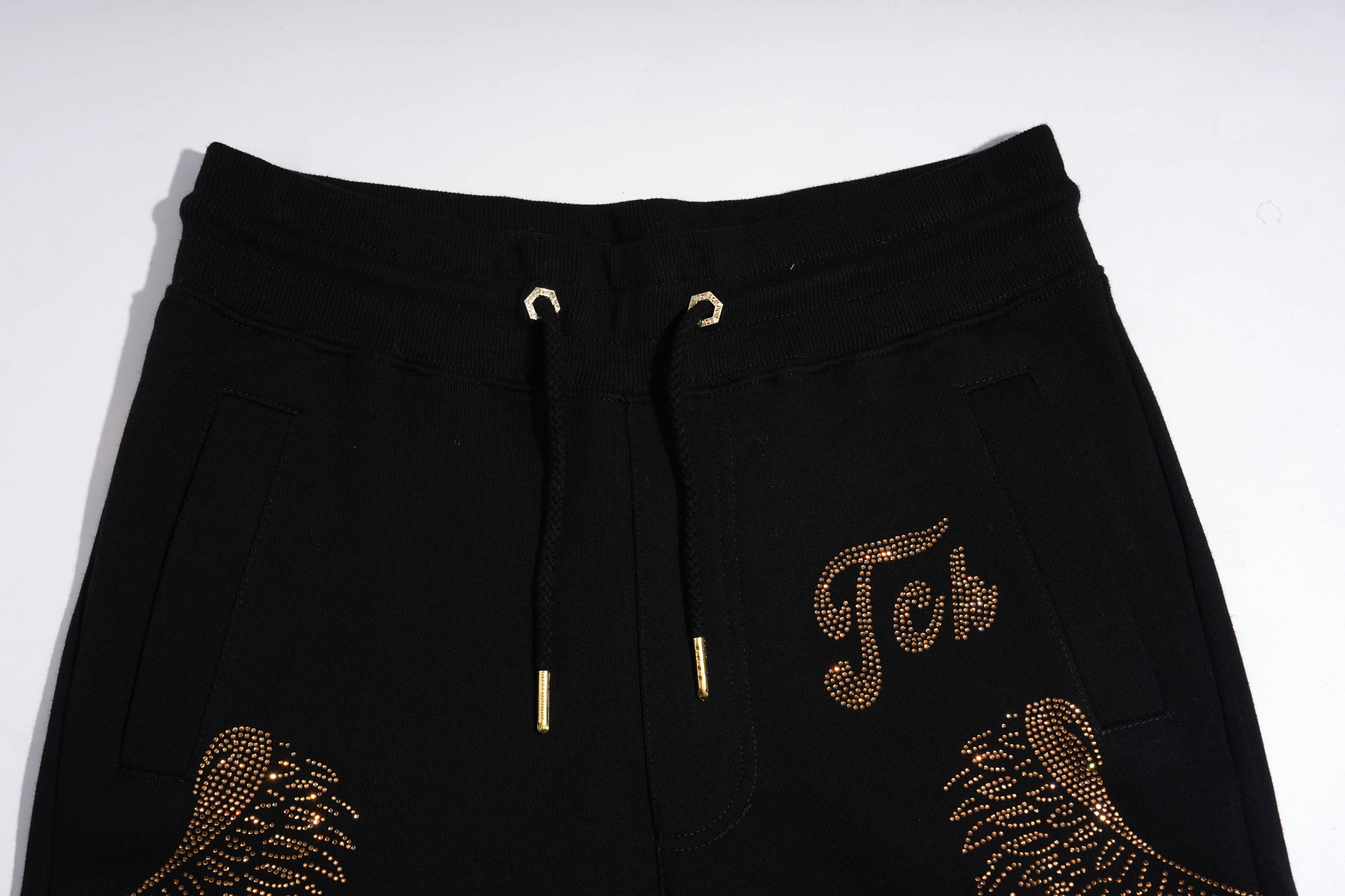 Black sweatpants featuring a gold rhinestone-embellished 'Joh' script on the left thigh, matching gold feather designs on the lower legs, and a black drawstring waist with gold metal tips. TCH Rhinestone Angel Wings Gold Detail Black Jogger Pants