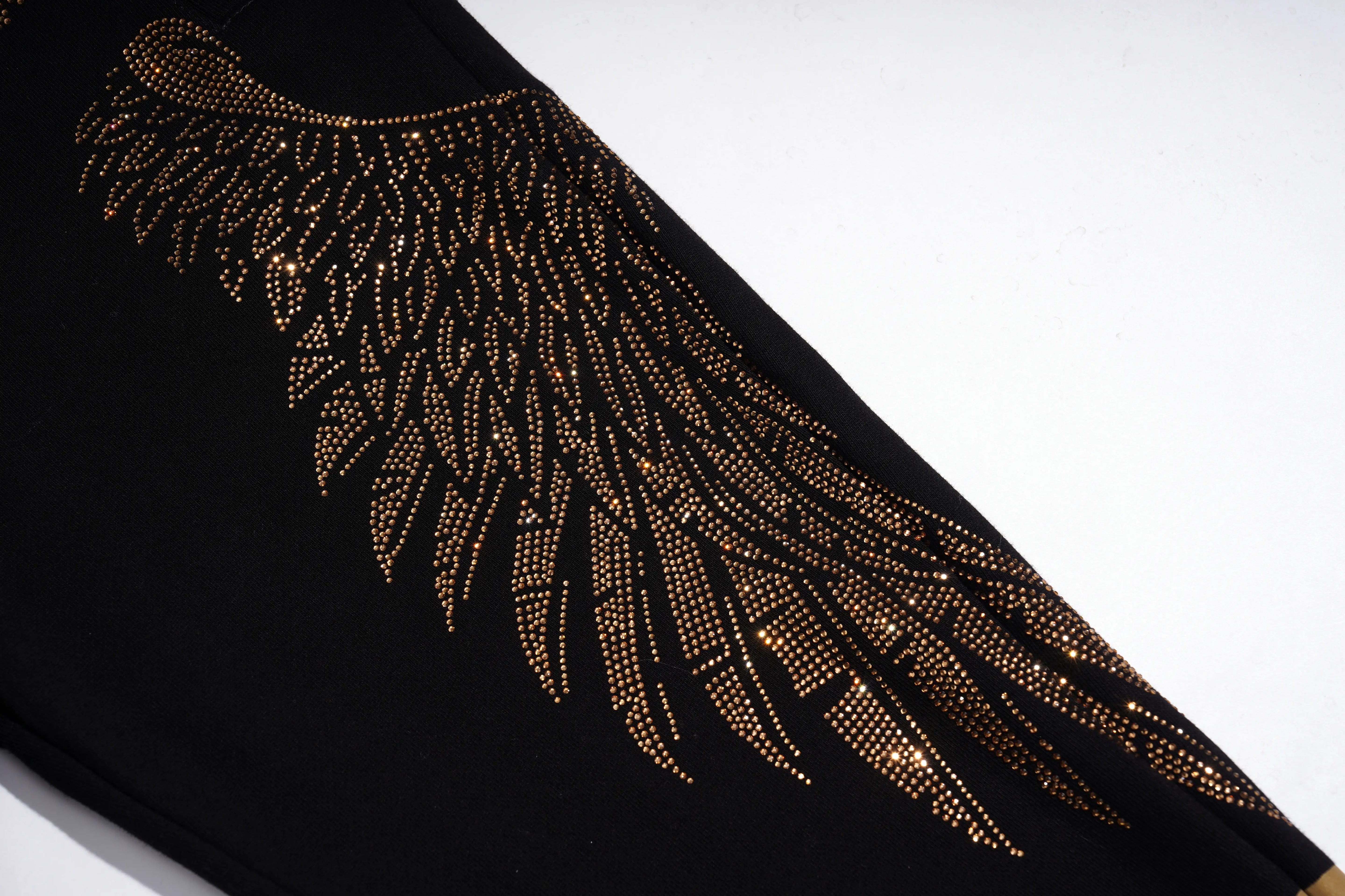 A close-up of a black garment featuring a large, detailed wing design embellished with shimmering gold rhinestones, set against a plain white background. TCH Rhinestone Angel Wings Gold Detail Black Jogger Pants