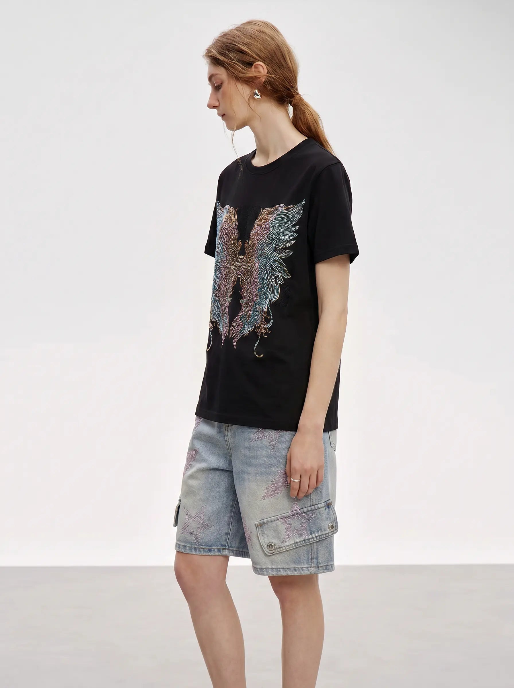 A woman modeling a black short-sleeve t-shirt with a large multicolor angel wing graphic on the front, paired with light-wash denim cargo shorts, set against a plain light gray studio background. TCH Rhinestone Angel Wings Graphic T-Shirt