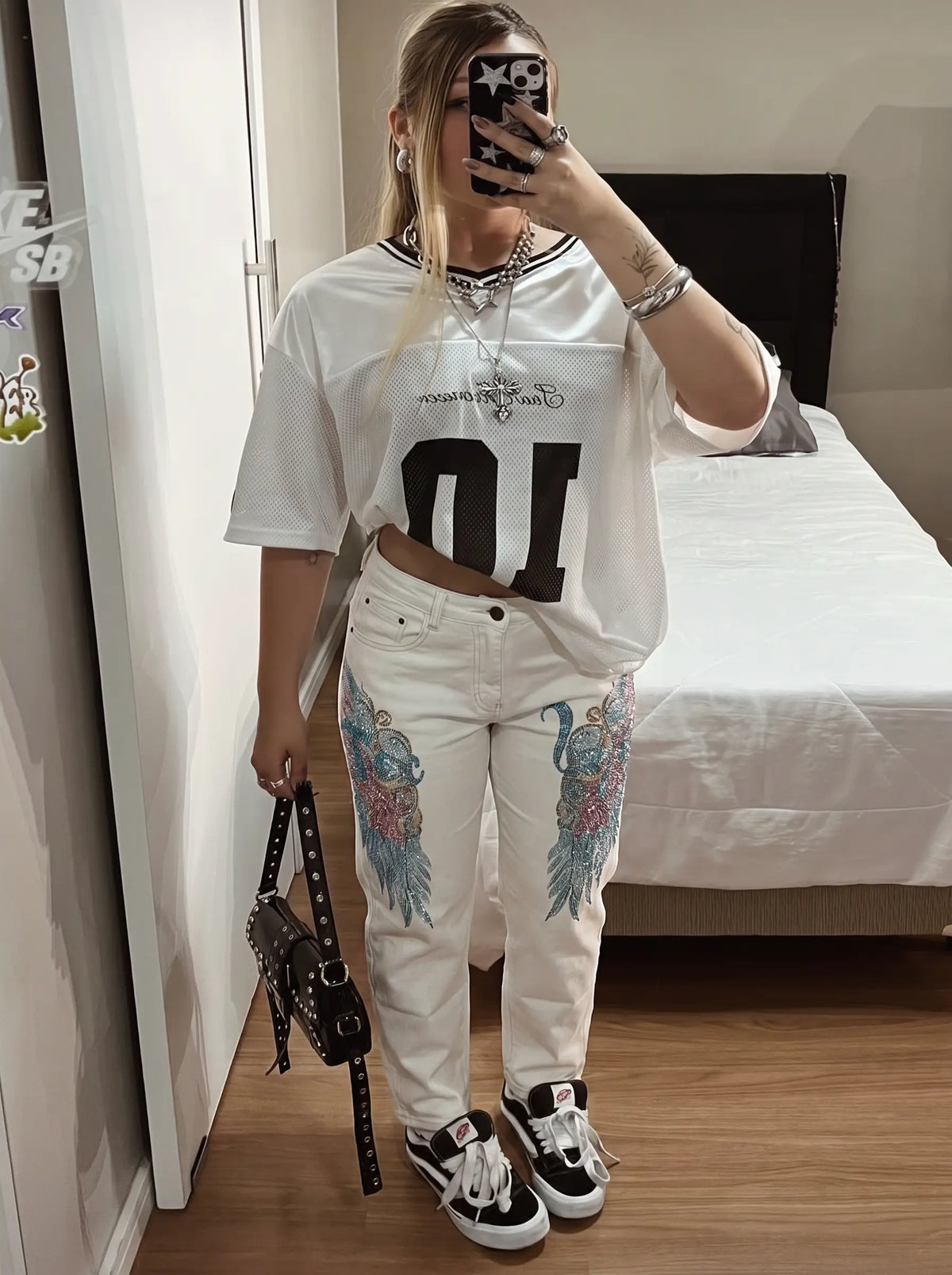 A woman takes a mirror selfie in a bedroom setting, wearing a white graphic t-shirt with a large black number '10' and 'Sparta FC' text, paired with white jeans featuring colorful wing designs on the thighs, and black and white sneakers. TCH Rhinestone Angel Wings White Straight-Leg Jeans