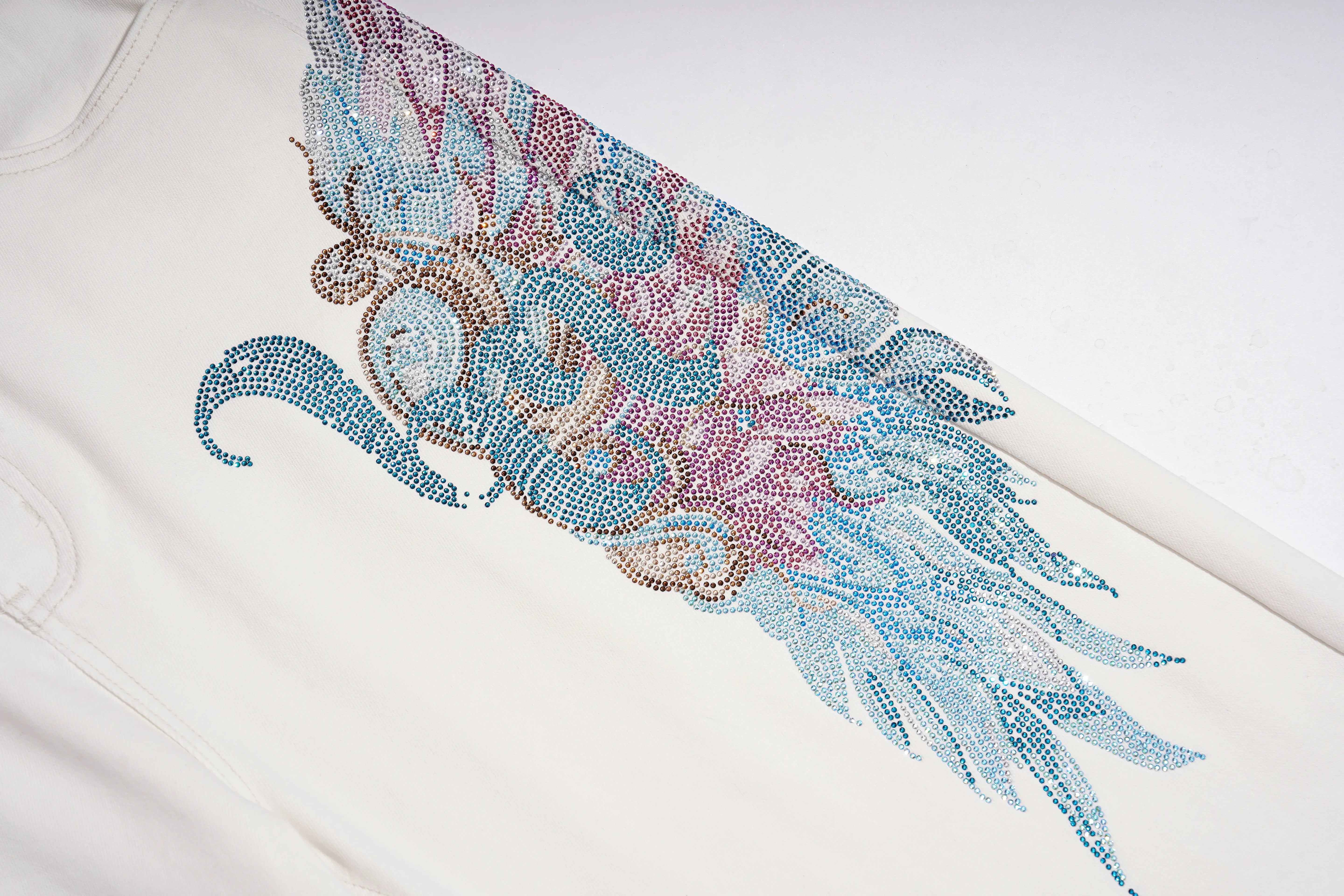 A close-up of a white garment featuring a large, detailed rhinestone wing design in shades of blue, pink, and gold. TCH Rhinestone Angel Wings White Straight-Leg Jeans