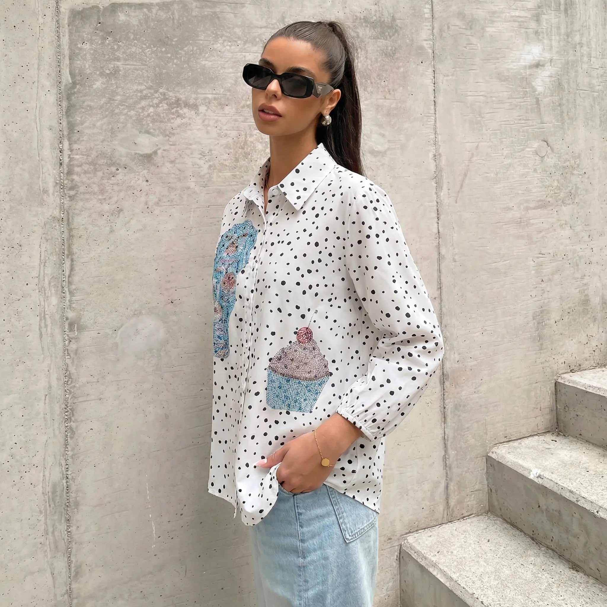 Woman wearing a white polka dot shirt with colorful cupcake designs, standing against a concrete wall. TCH Rhinestone Animal Polka Dot Shirt